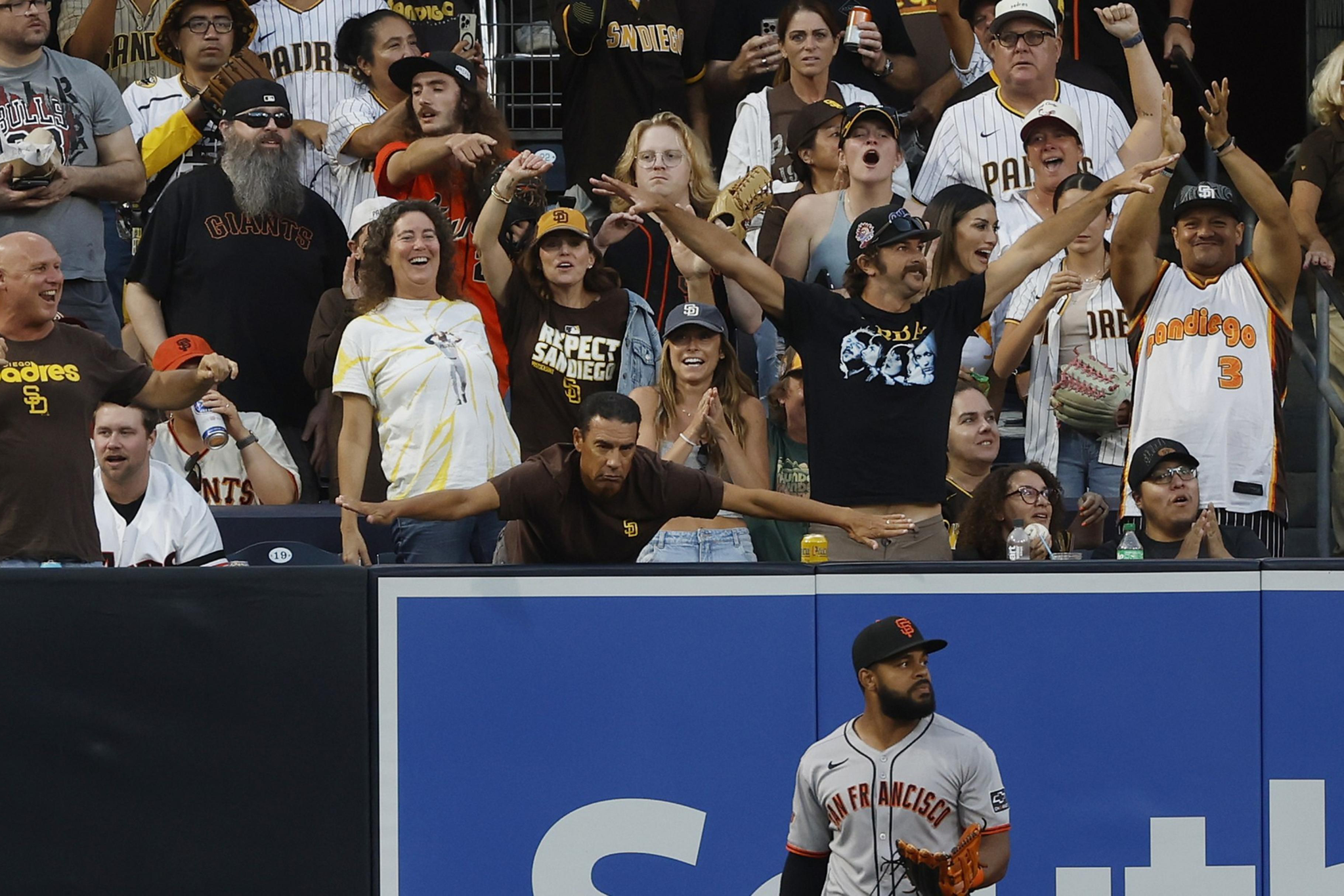 A San Francisco Giants player stands near the outfield wall as energetic fans behind him, mostly wearing San Diego Padres gear, cheer and react.