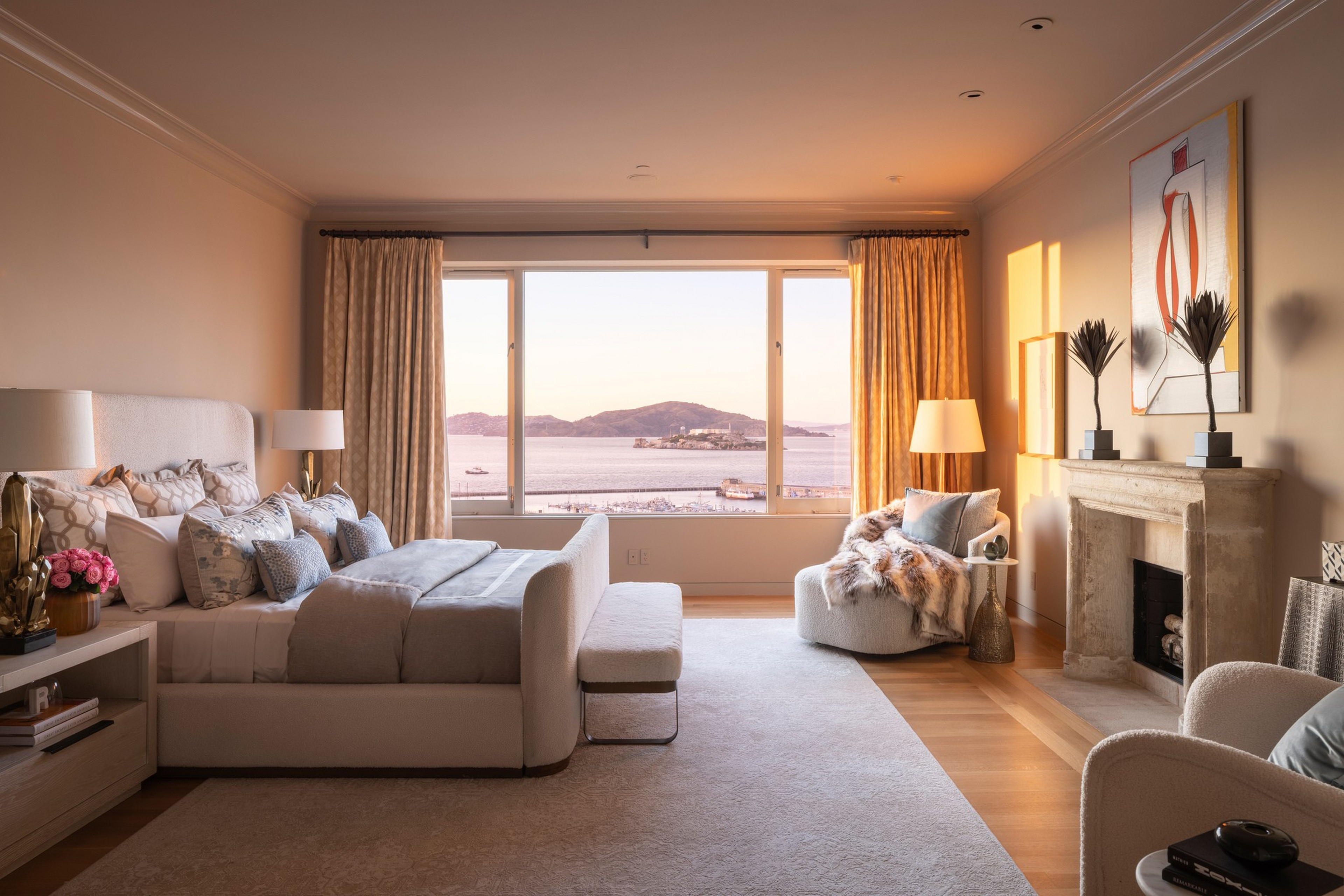 A cozy bedroom with a large bed, soft pillows, a sitting area, a fireplace, elegant curtains, and a big window showing a serene ocean view with islands.