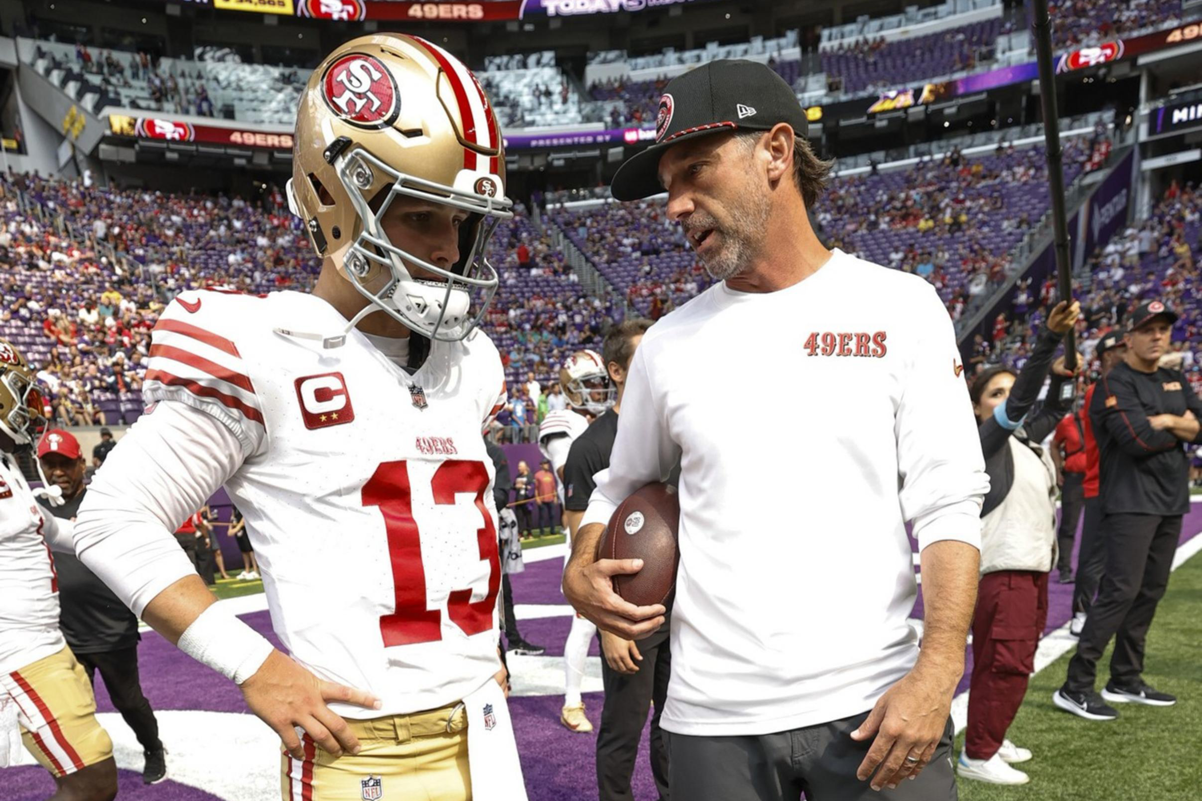 A football player in a white and gold uniform stands on the sideline with a man in a white 49ers shirt holding a football, talking together on a stadium field.