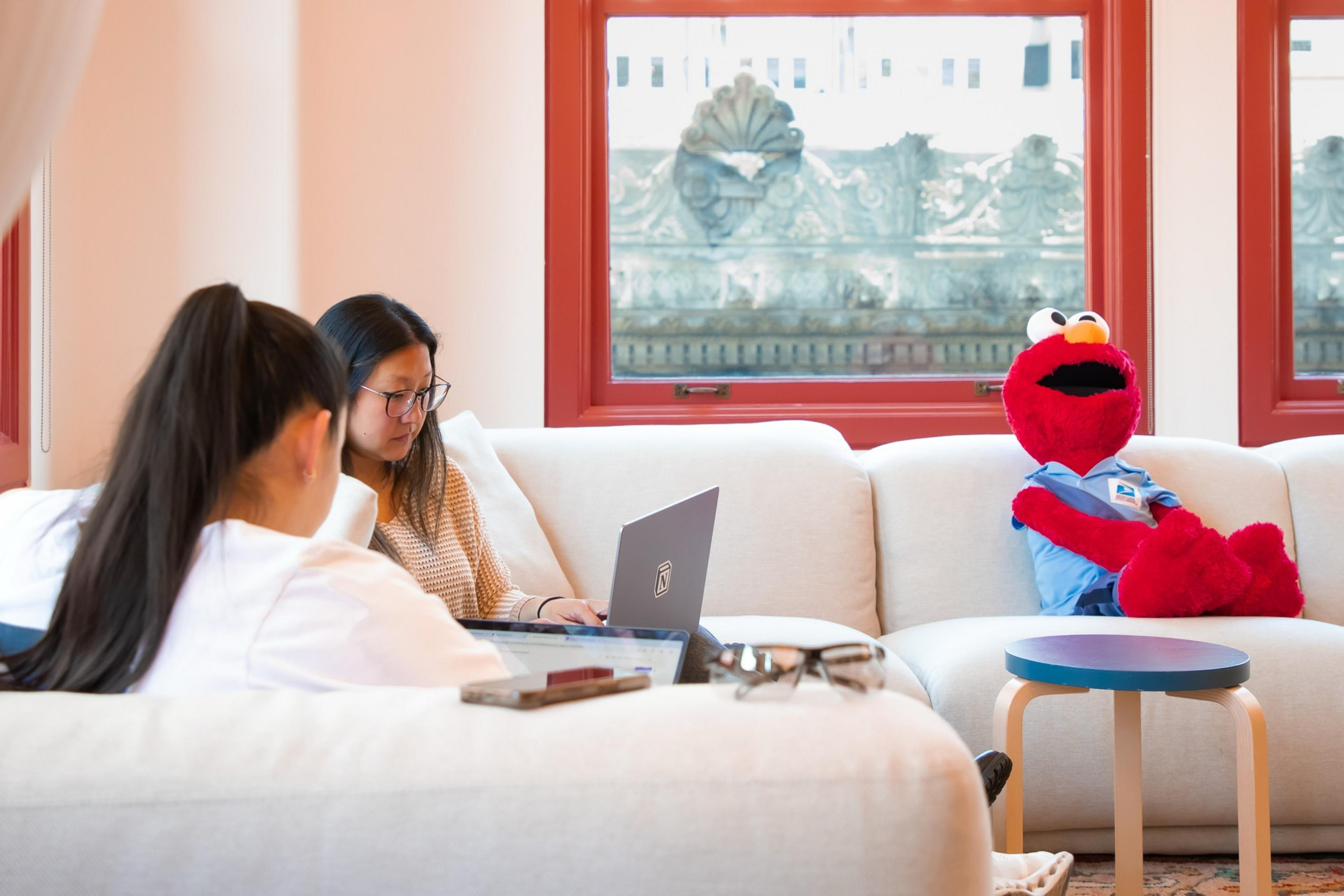 Two people are on a white sofa using laptops, with a red Muppet character in a blue shirt sitting beside them, framed by red windows.