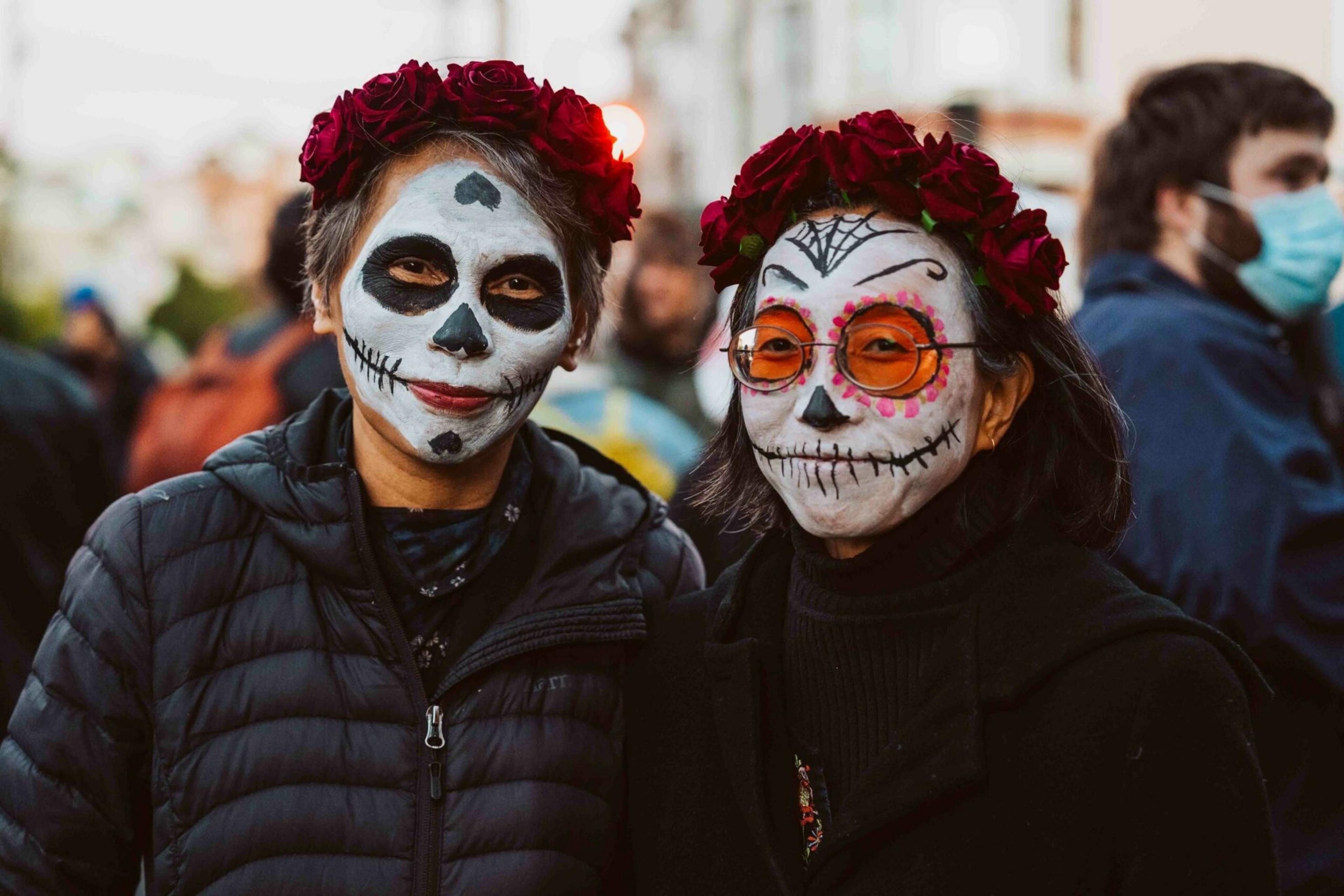 Two people with skull face paint and red flower crowns pose together outdoors during a festive event, with other masked individuals in the background.
