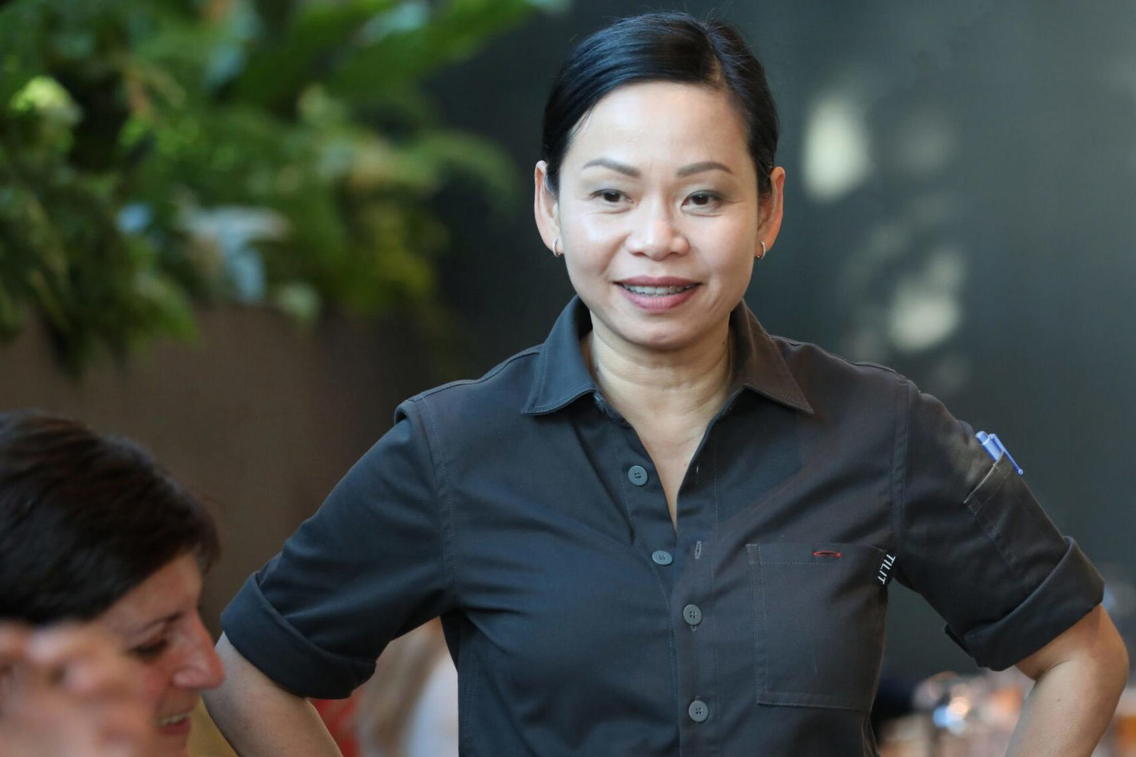 A woman in a gray uniform stands with hands on hips, smiling in a restaurant setting with people seated at a long table. There are plants and table settings visible.