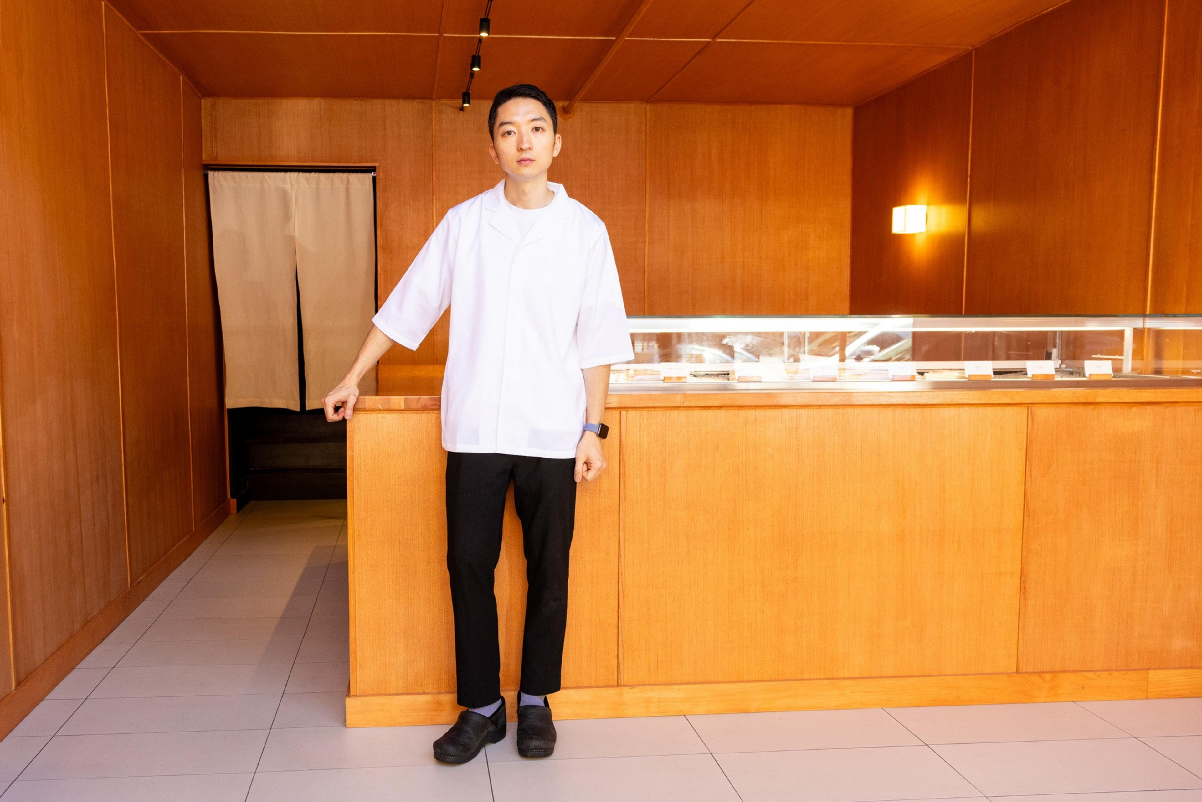 A man in a white coat stands behind a wooden counter in a minimalistic, warmly lit room, with beige curtains in the background and a display case on the counter.