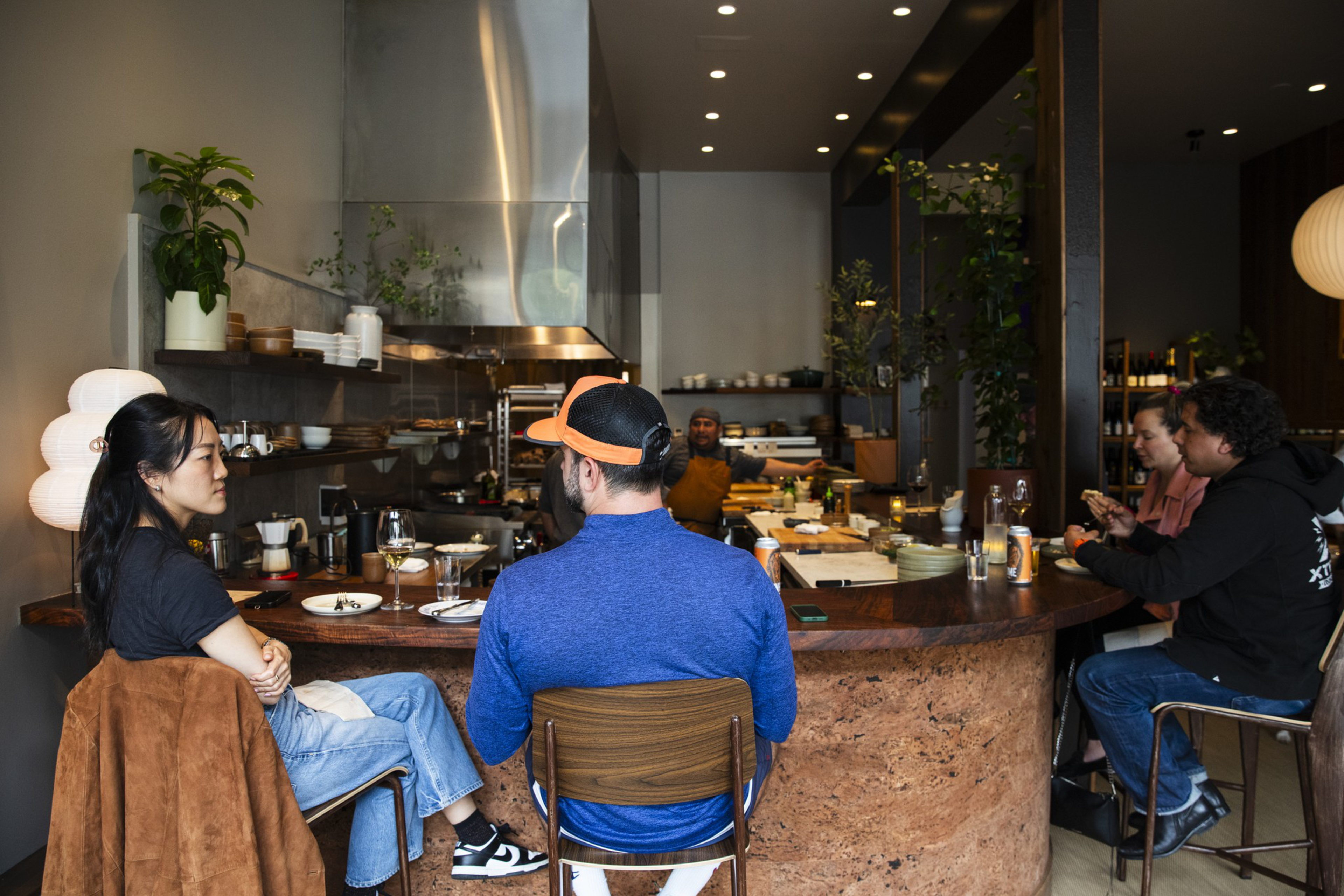 Five people sit at a curved counter in a modern kitchen, engaged in conversation and dining, with shelves, plants, and kitchen tools in the background.
