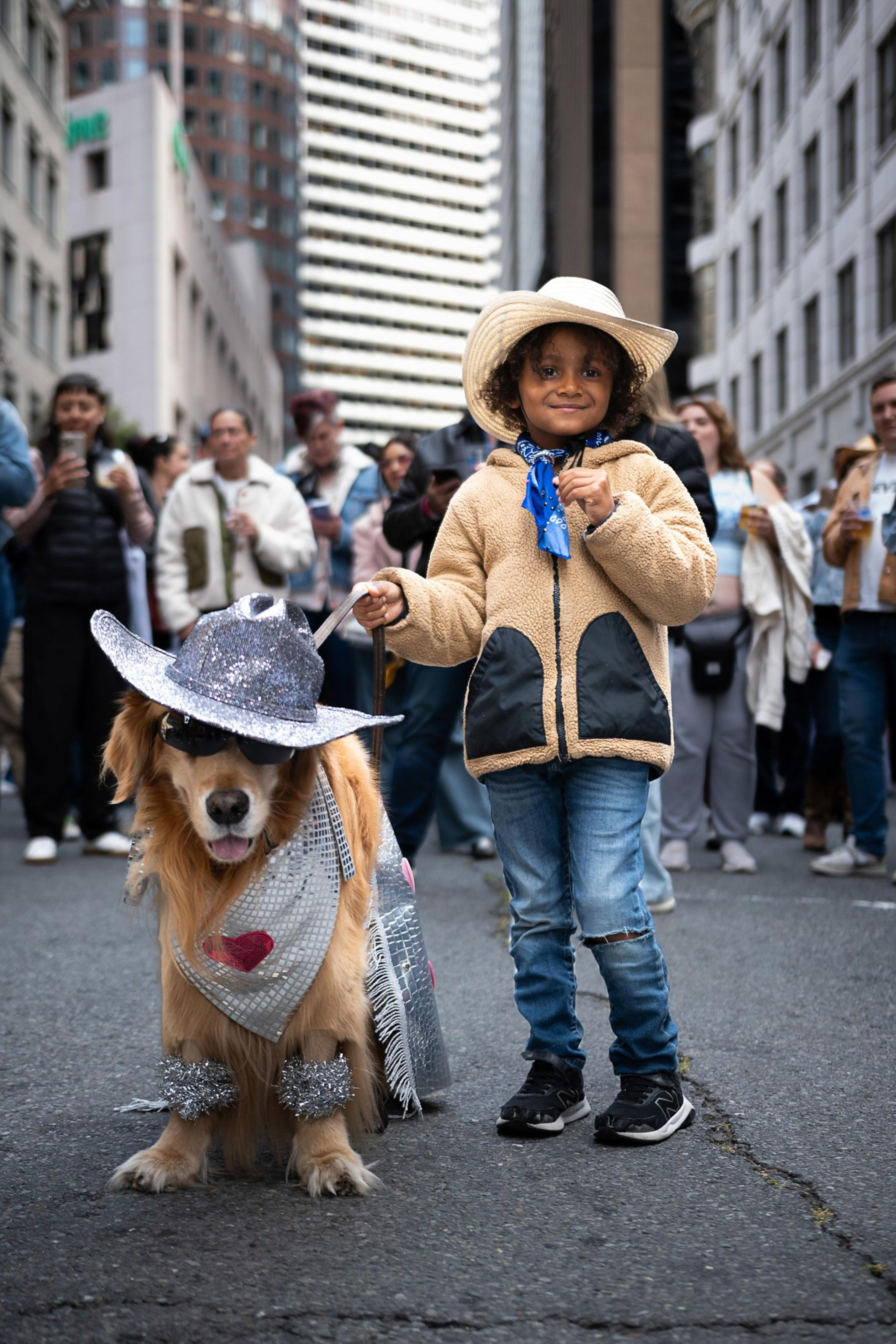 A child in a cowboy hat stands next to a dog dressed in sparkly cowboy attire. They are on a city street, surrounded by smiling onlookers.