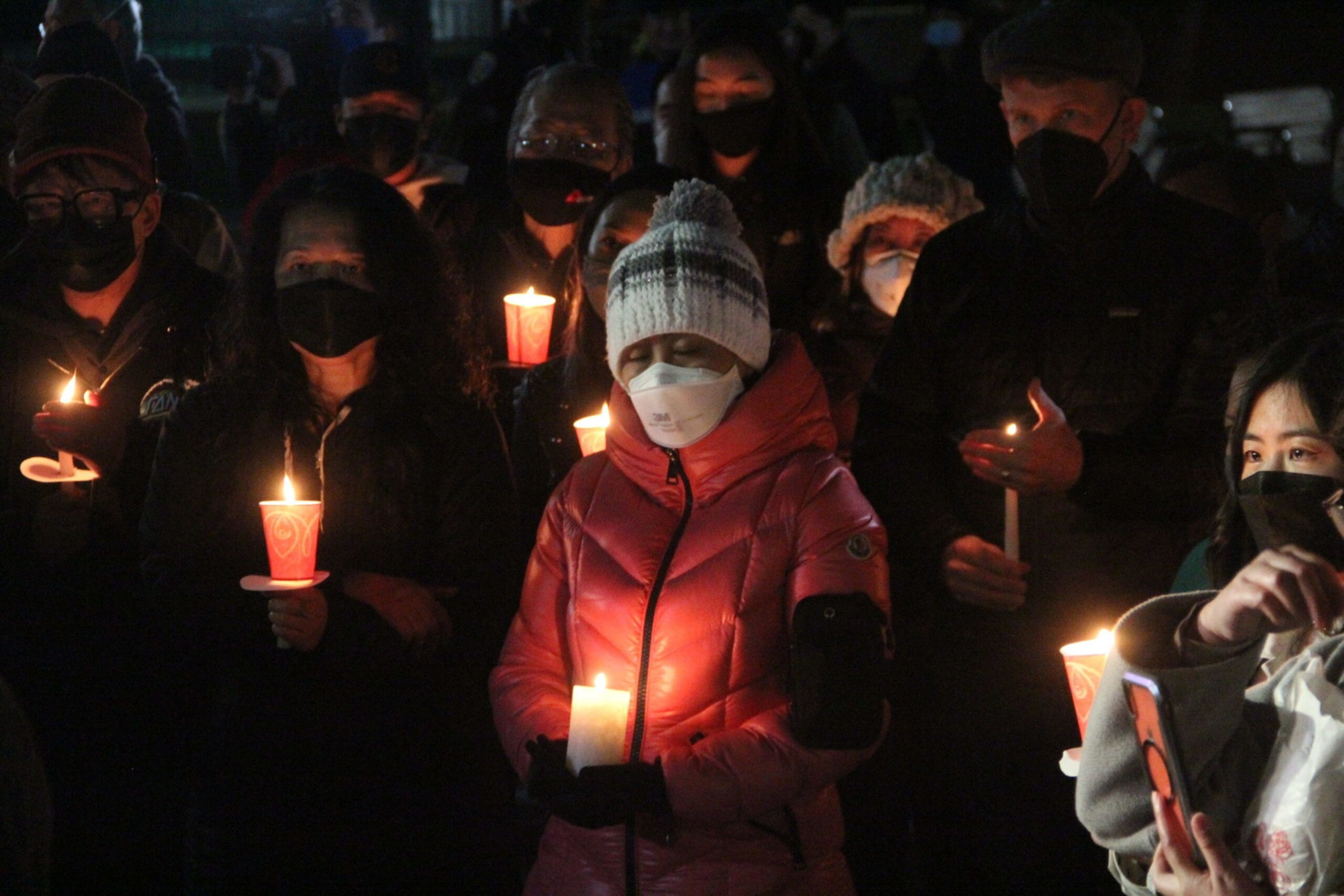 A group of people wearing masks hold lit candles during a nighttime vigil, with one woman in a pink jacket and white knit hat in the center.