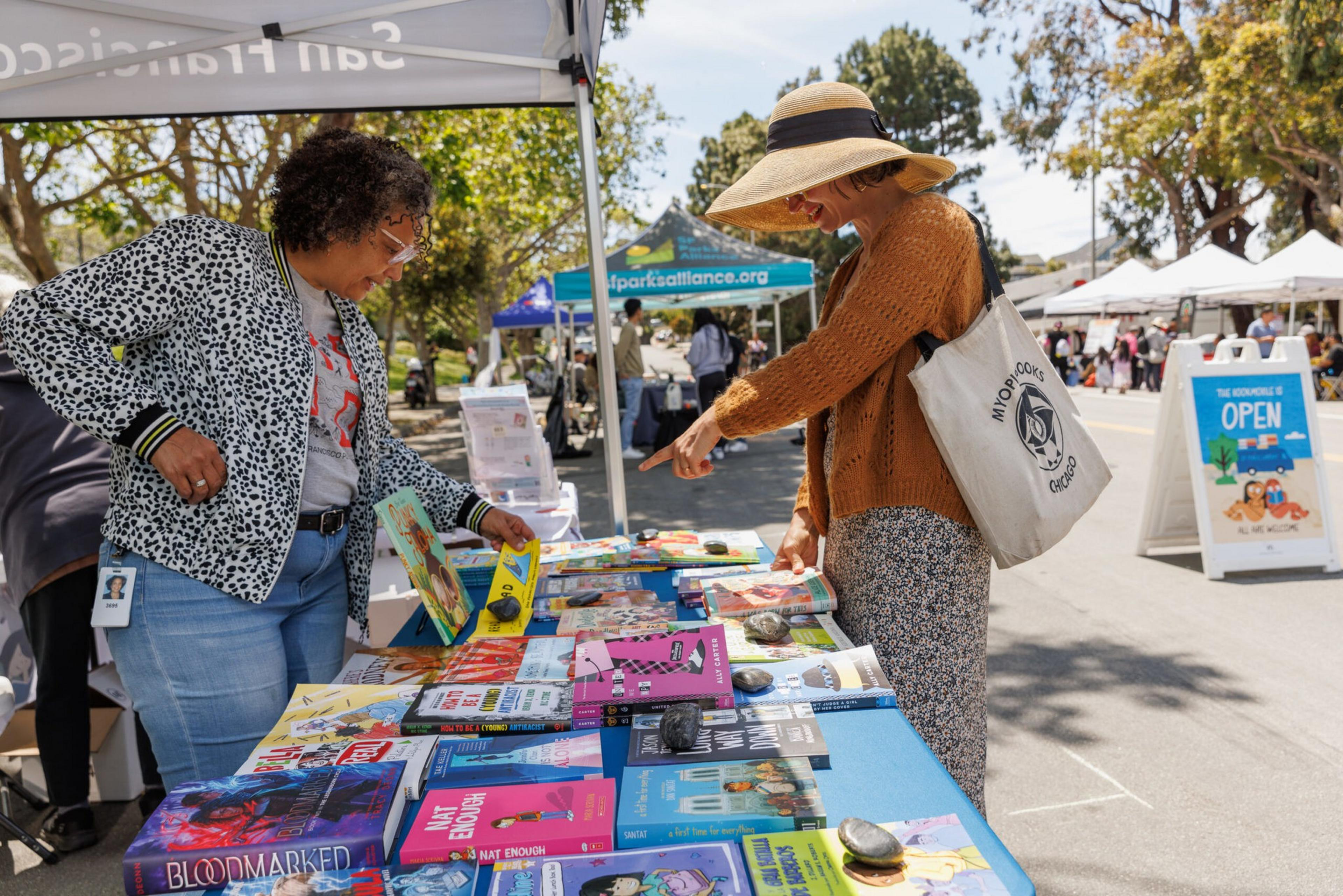 Two women browse books at an outdoor stall under a sunny sky.