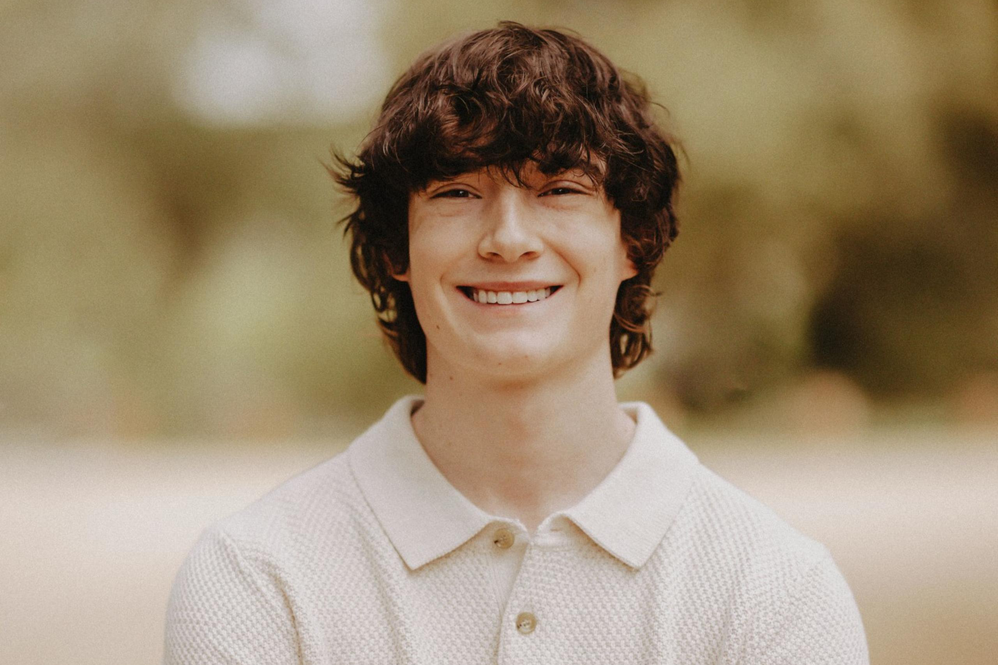 A young man with curly brown hair smiles warmly, wearing a light beige short-sleeve polo shirt, standing with arms crossed in a blurred outdoor setting.
