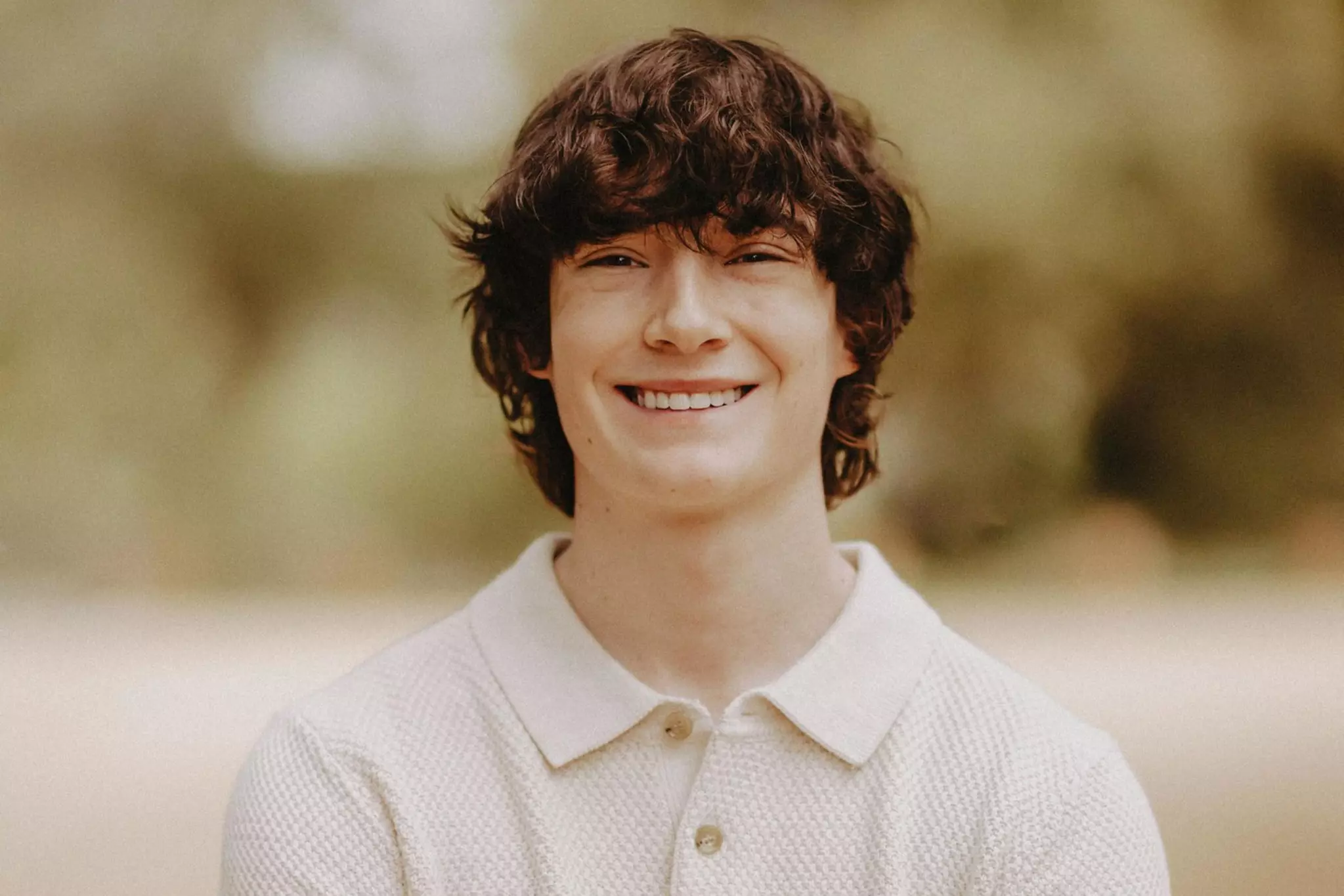 A young man with curly brown hair smiles warmly, wearing a light beige short-sleeve polo shirt, standing with arms crossed in a blurred outdoor setting.