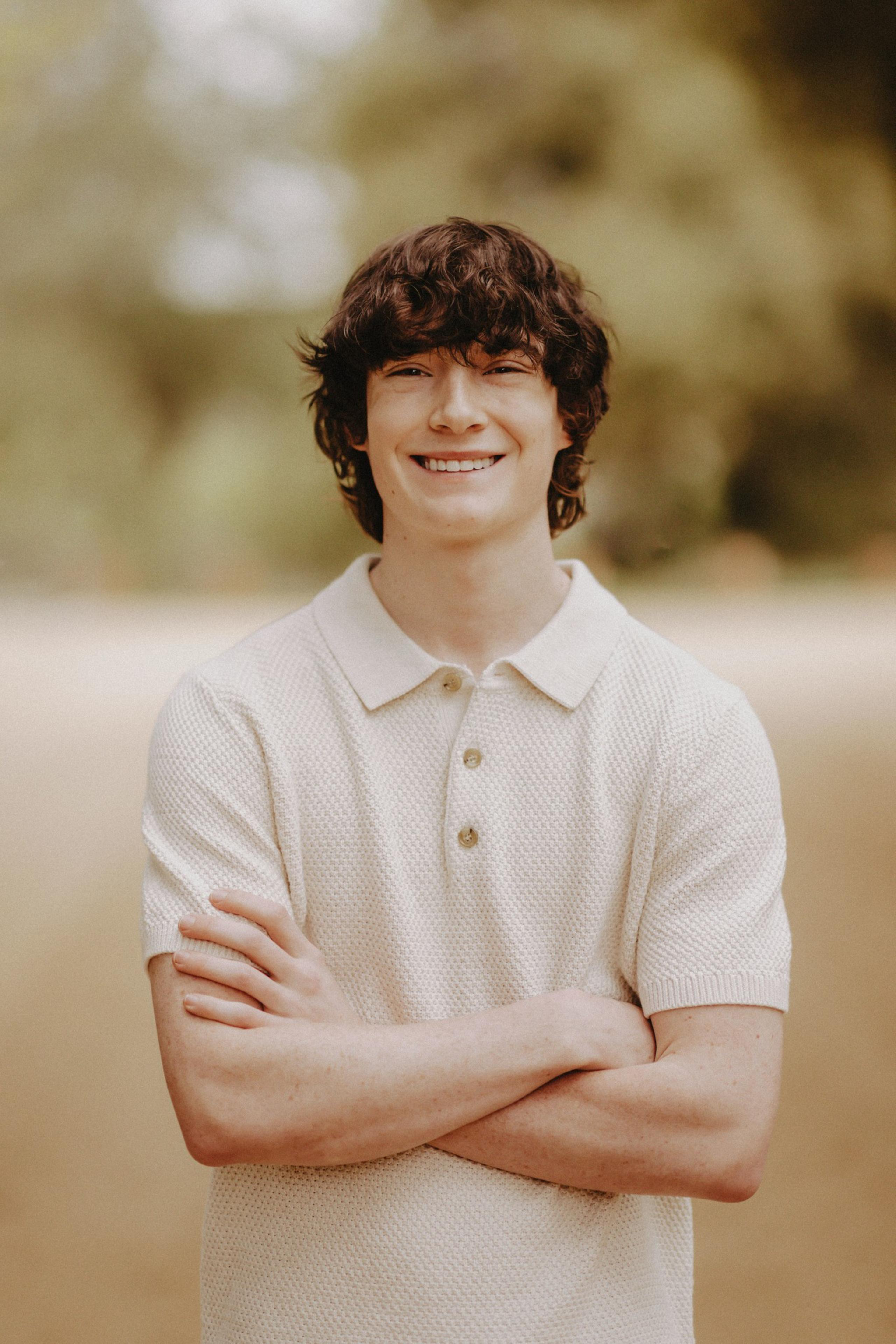 A young man with curly brown hair smiles warmly, wearing a light beige short-sleeve polo shirt, standing with arms crossed in a blurred outdoor setting.
