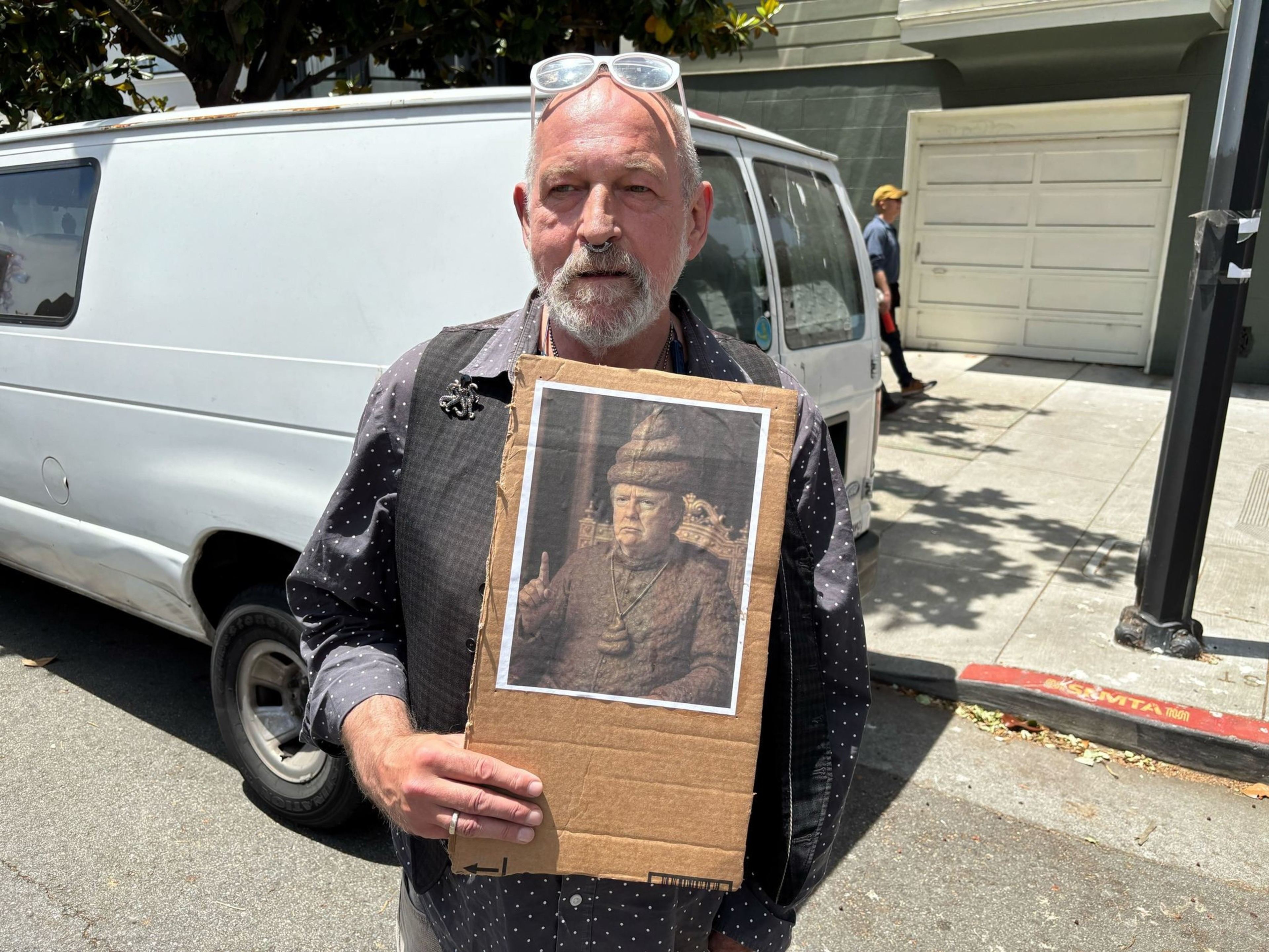 A man stands outdoors holding a cardboard with an image of a historical figure. He's in front of a white van and wearing sunglasses on his head.