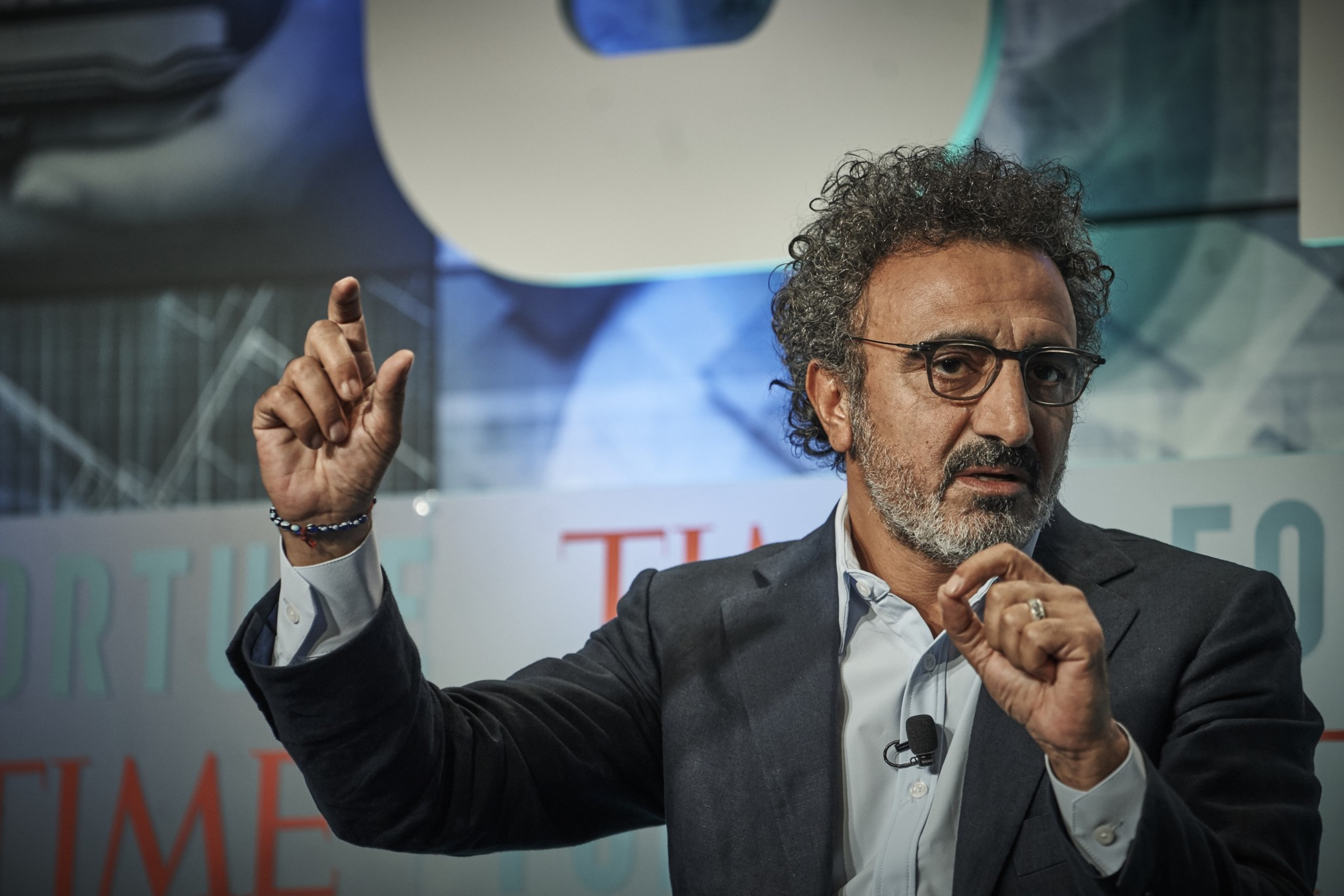 A man with curly hair and glasses, wearing a suit, gestures while speaking in front of a blurred, colorful background with text.