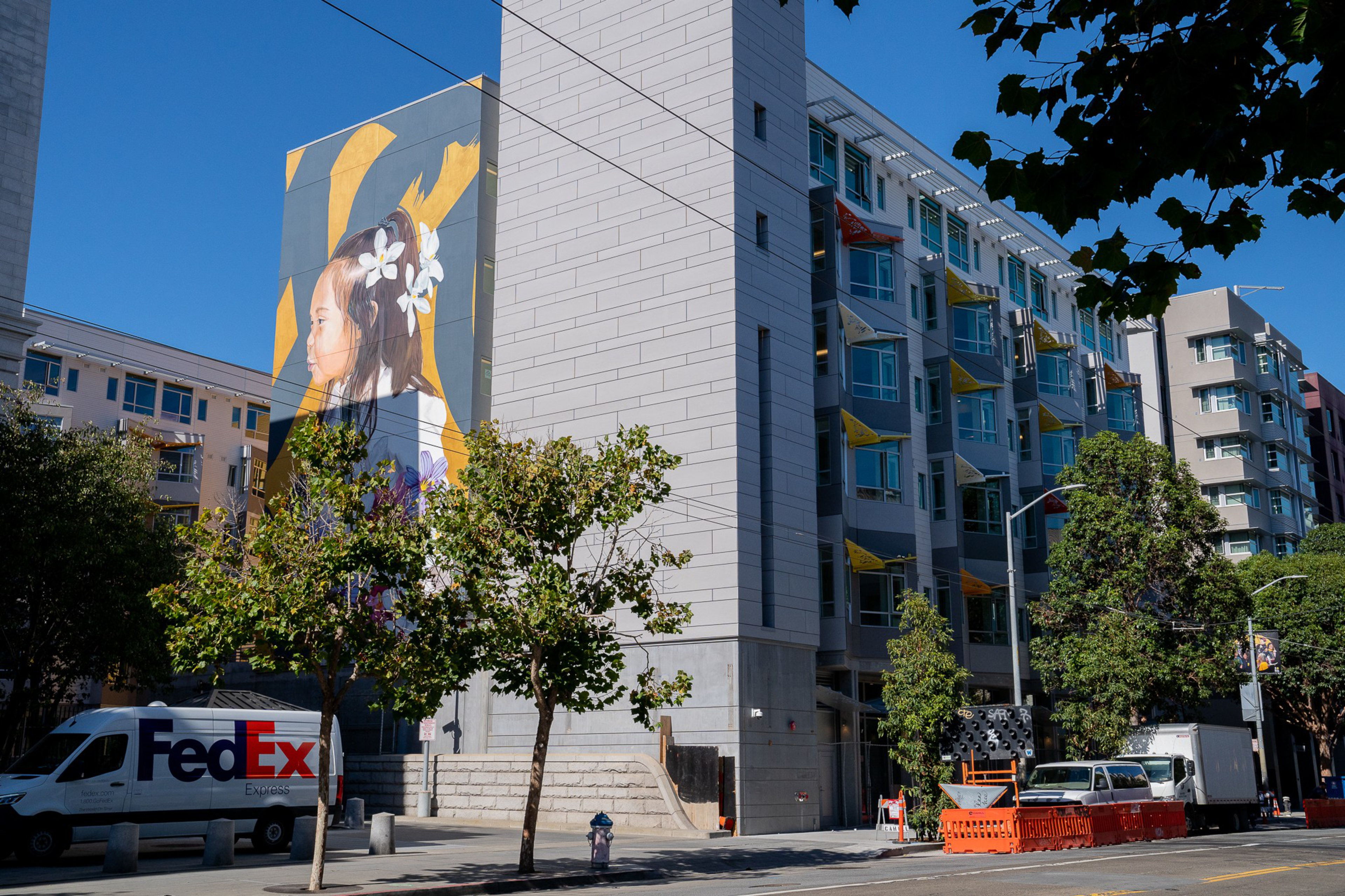 A large mural of a girl with flowers in her hair decorates the side of a modern apartment building under a clear blue sky.