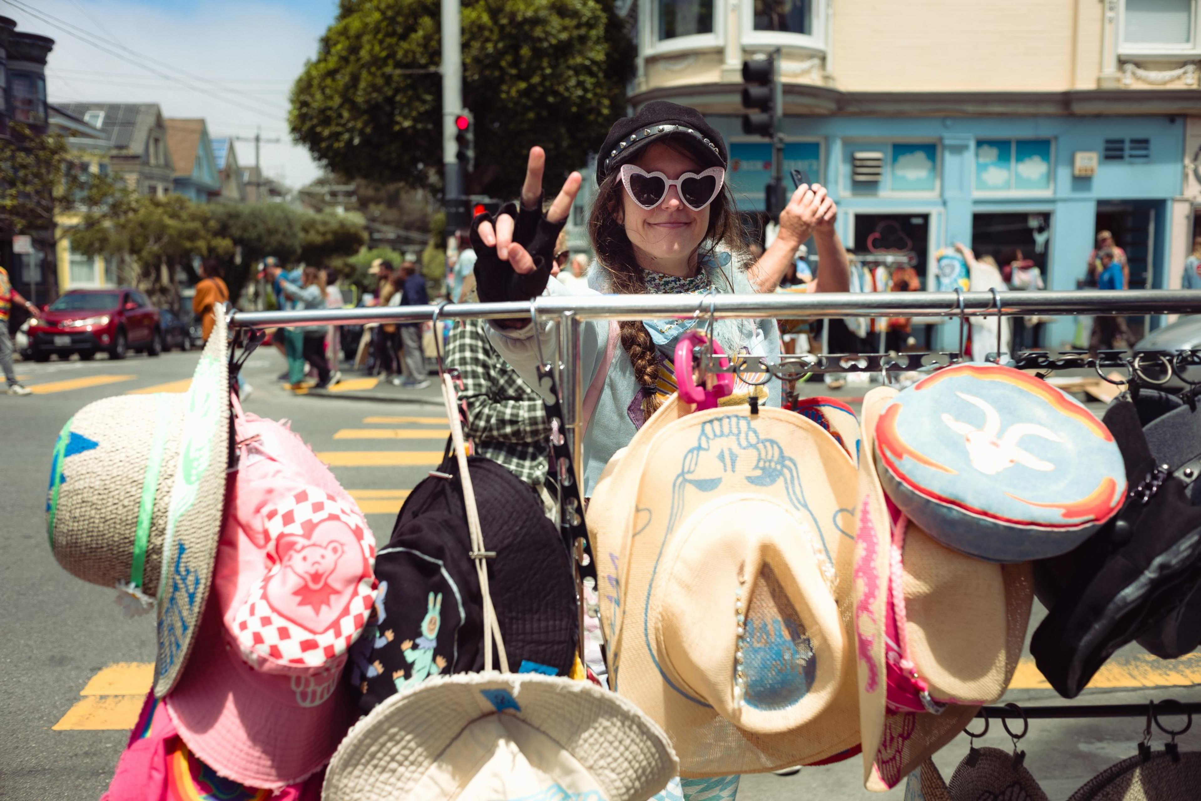 A smiling person wearing heart-shaped sunglasses and a black cap flashes a peace sign behind a rack of colorful hats on a sunny street.