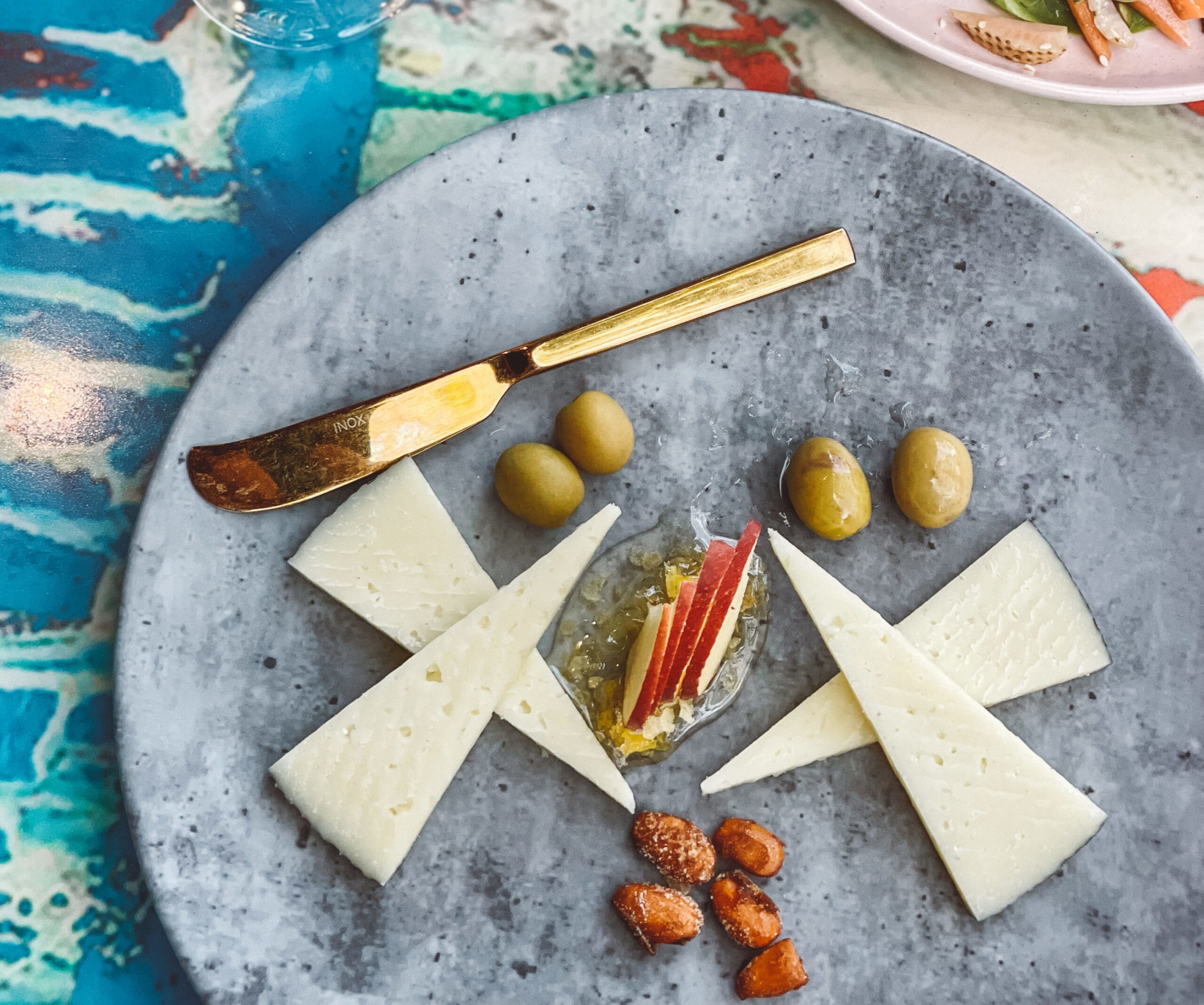 Two sets of triangular cheese slices, green olives, honey with apple slices, and a few almonds are arranged on a gray plate with a small gold spreader.