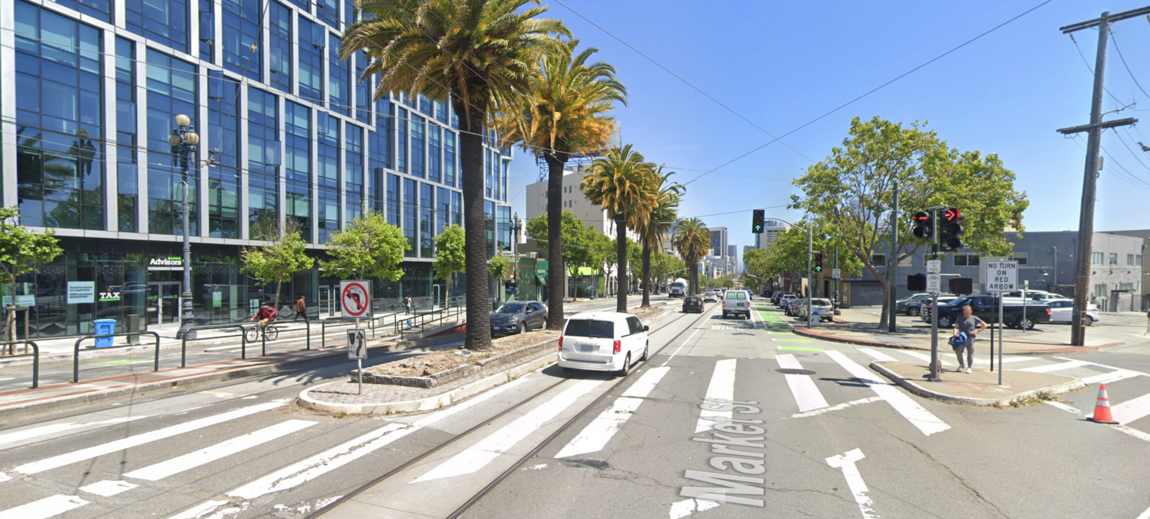 A city street with palm trees, crosswalks, traffic lights, cars, a bike lane, and modern glass buildings lining the road under clear skies.