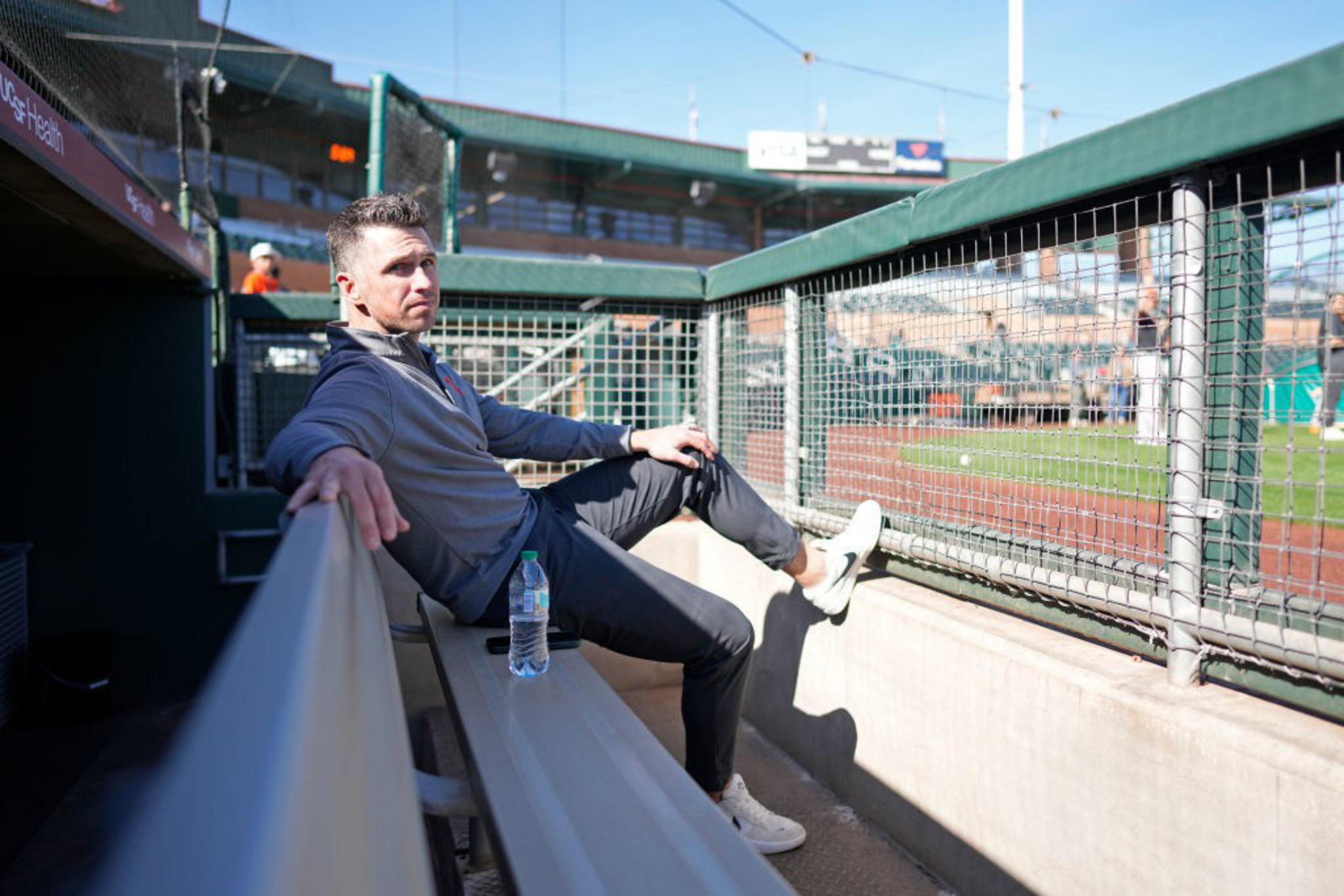 A man sits in a baseball dugout, relaxed with legs crossed. He's wearing a gray jacket and black pants. A water bottle rests on the bench beside him.