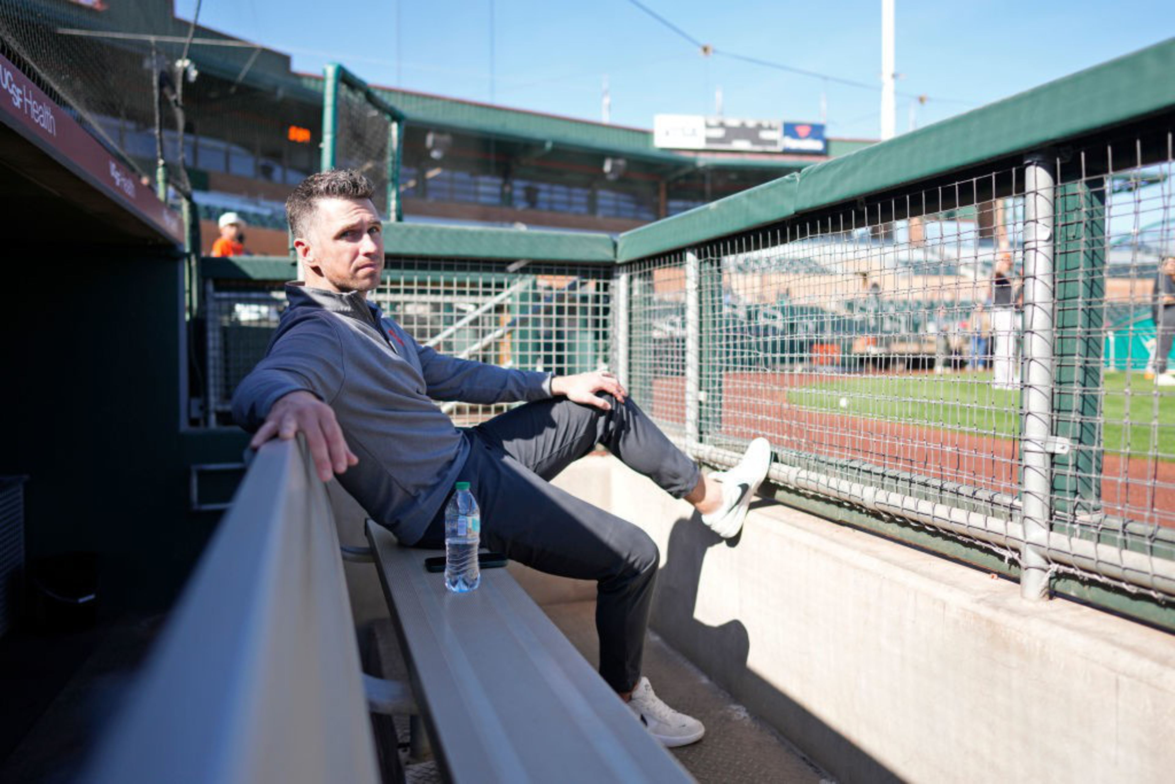 A man sits in a baseball dugout, relaxed with legs crossed. He's wearing a gray jacket and black pants. A water bottle rests on the bench beside him.