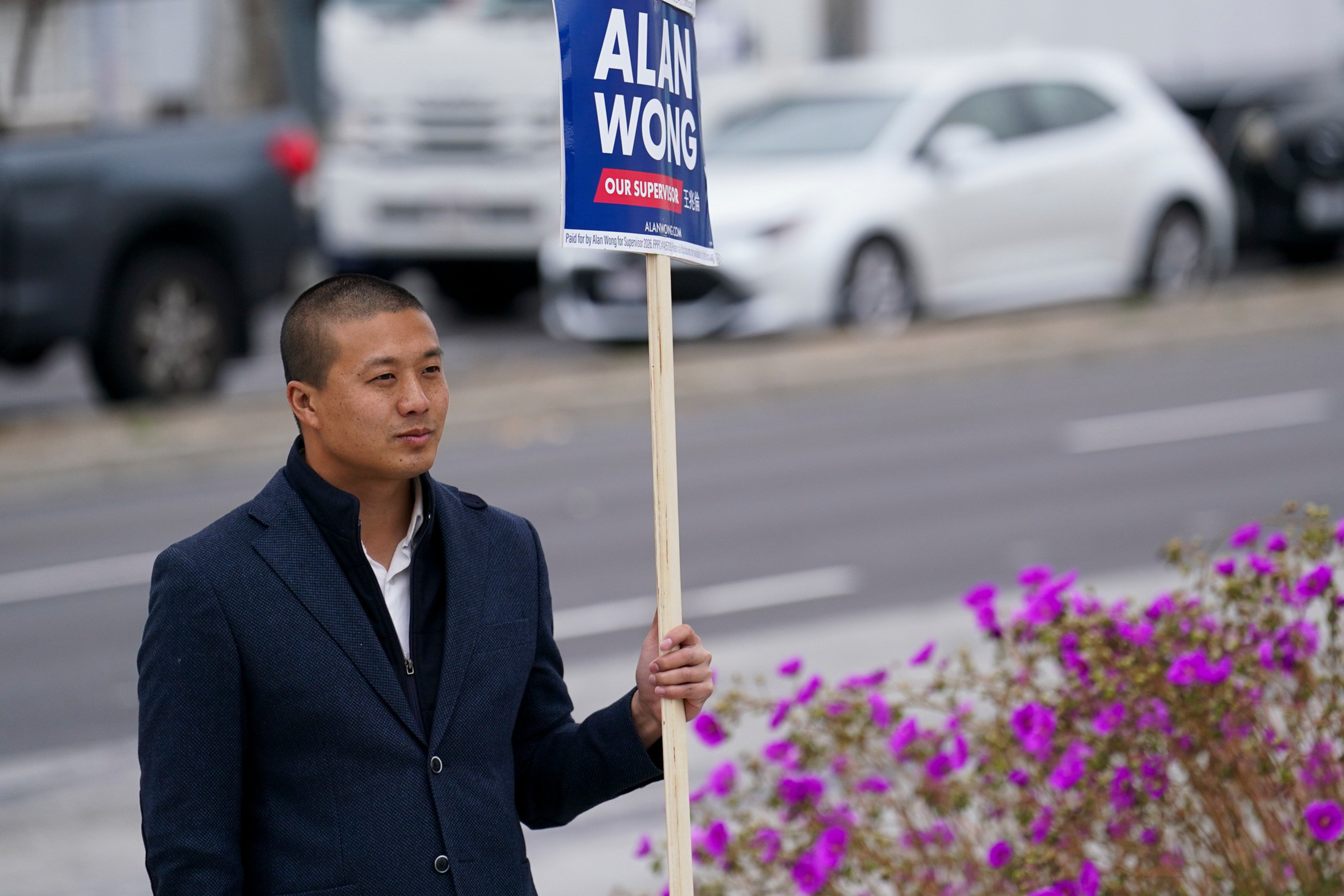 A man in a dark jacket stands by a roadside holding a campaign sign that reads "Alan Wong Our Supervisor," with purple flowers nearby.