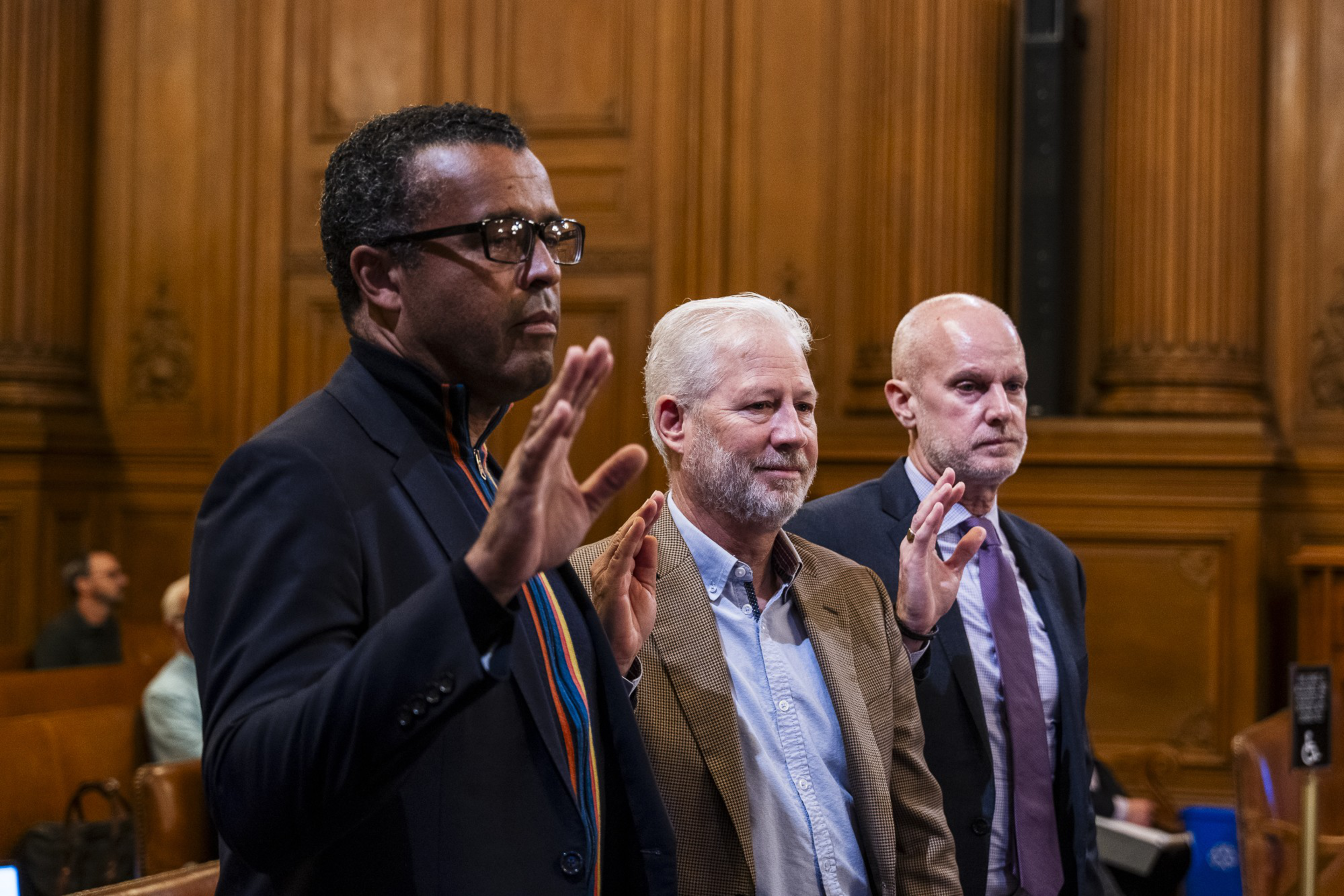 Three men are standing with raised hands in a wood-paneled room, possibly taking an oath. They are wearing formal attire and appear serious.