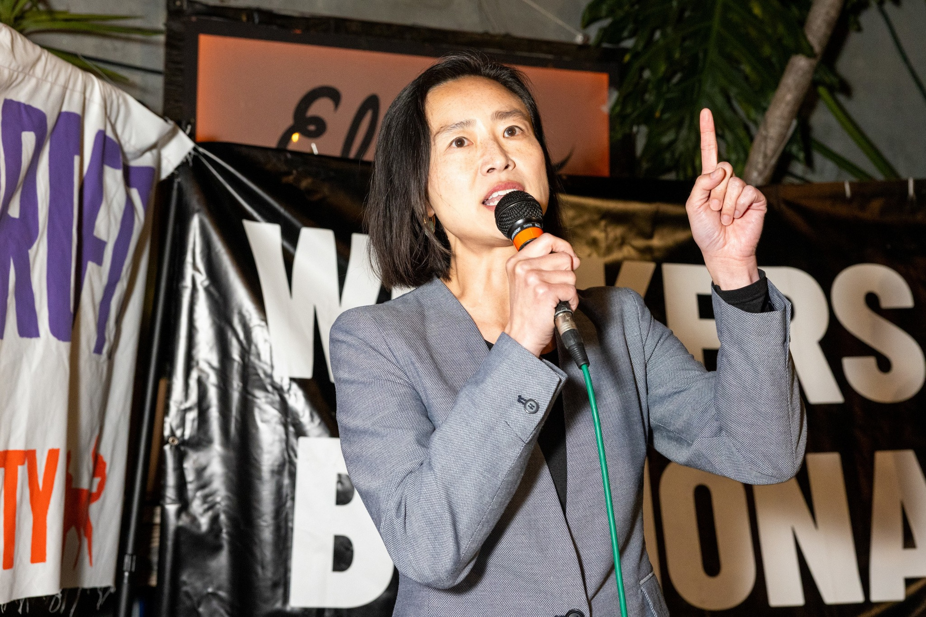 A person in a gray blazer speaks into a microphone, raising one finger, standing in front of black and white protest banners.