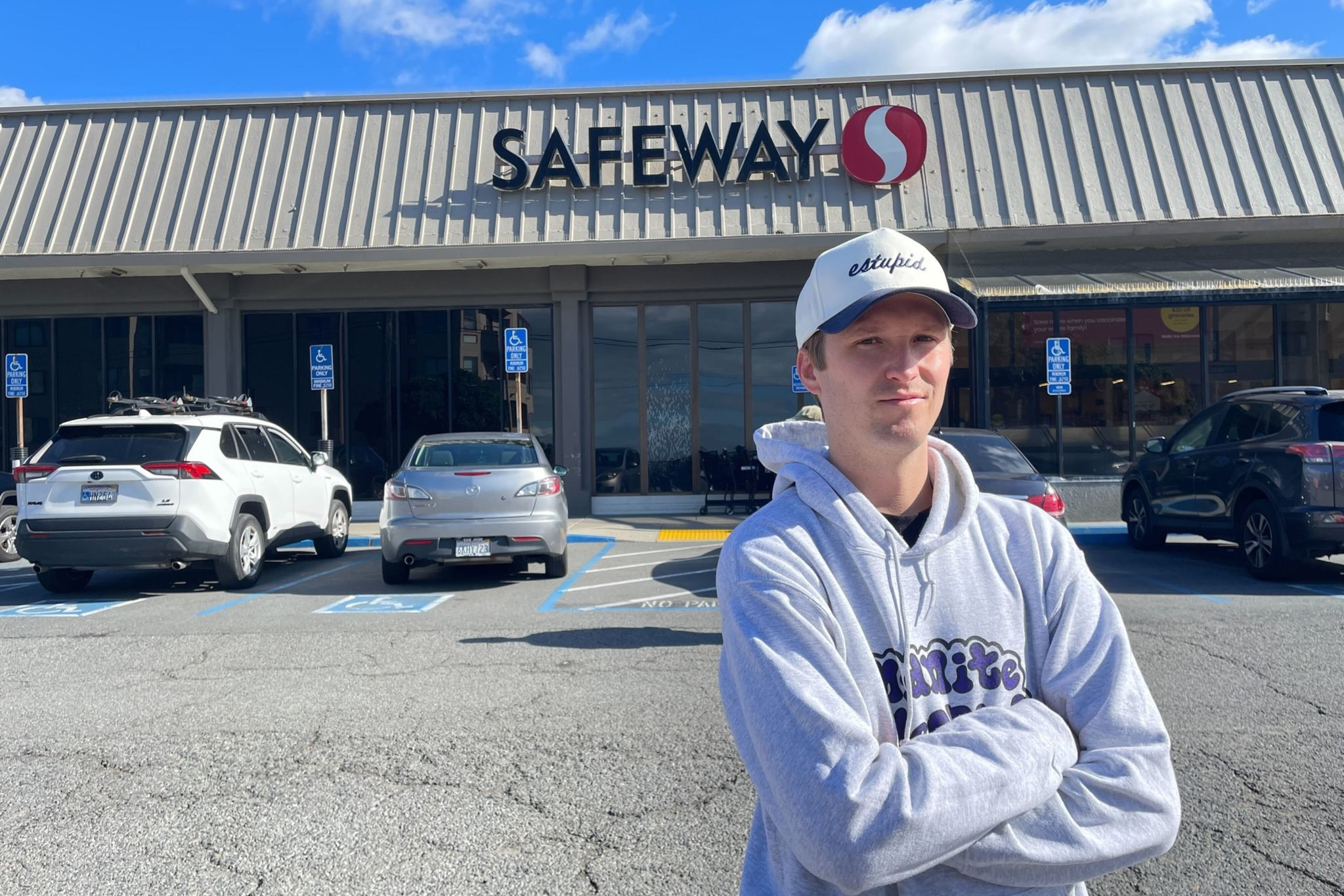 A man wearing a light gray hoodie and white cap stands with arms crossed in front of a Safeway store parking lot on a sunny day.