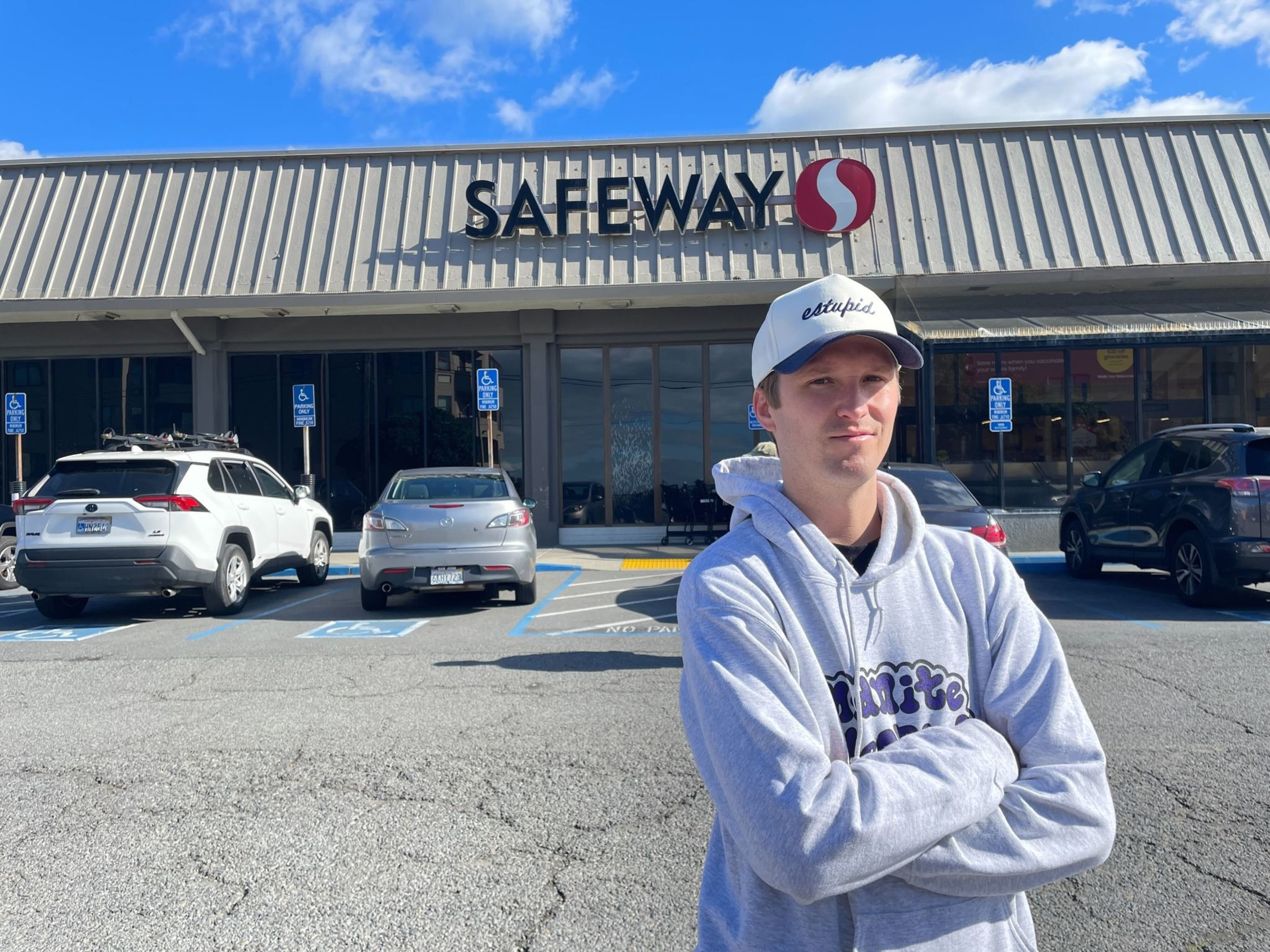 A man wearing a light gray hoodie and white cap stands with arms crossed in front of a Safeway store parking lot on a sunny day.