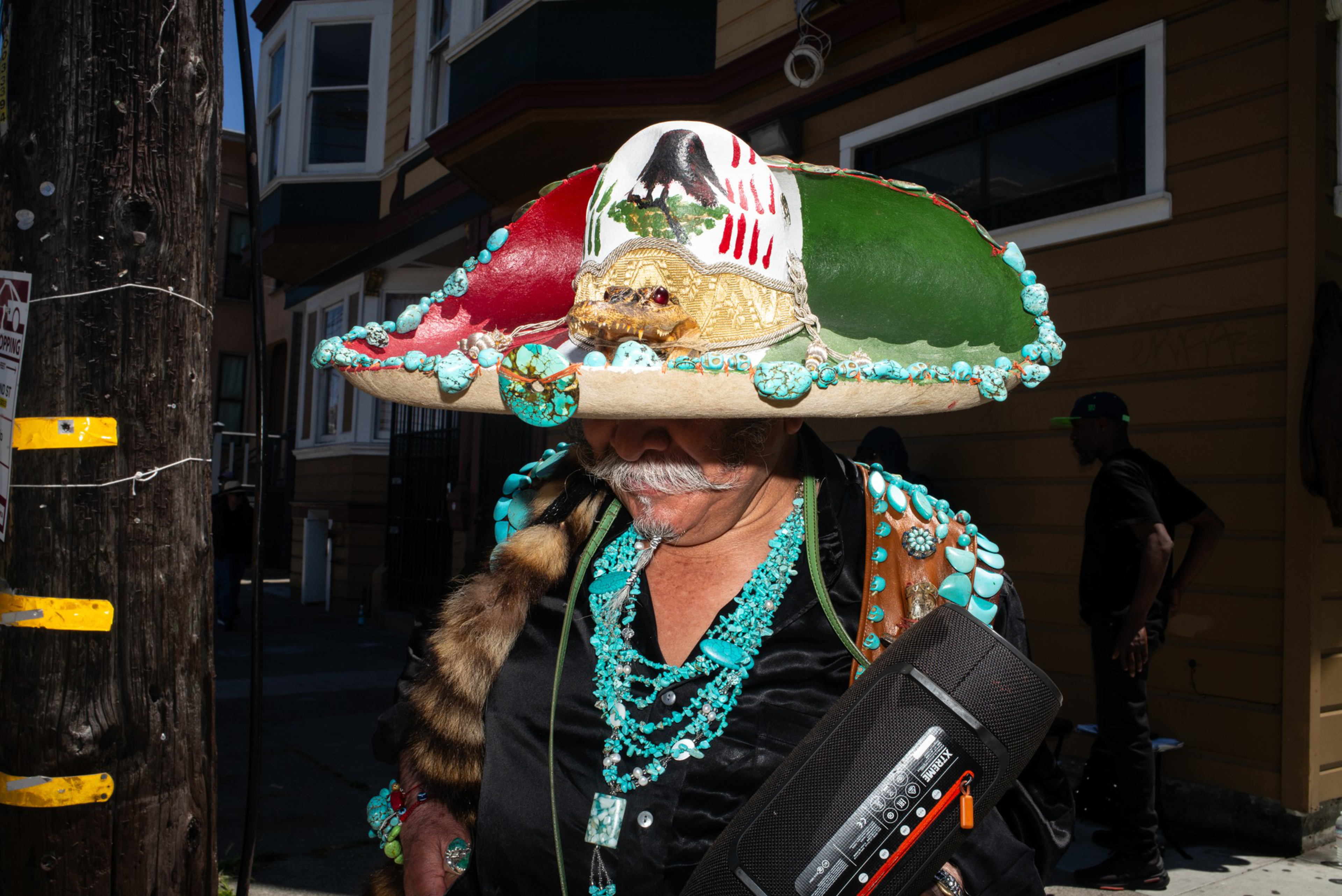 A man wears a large sombrero decorated with a painted skull and turquoise stones, a turquoise necklace, and carries a black case.