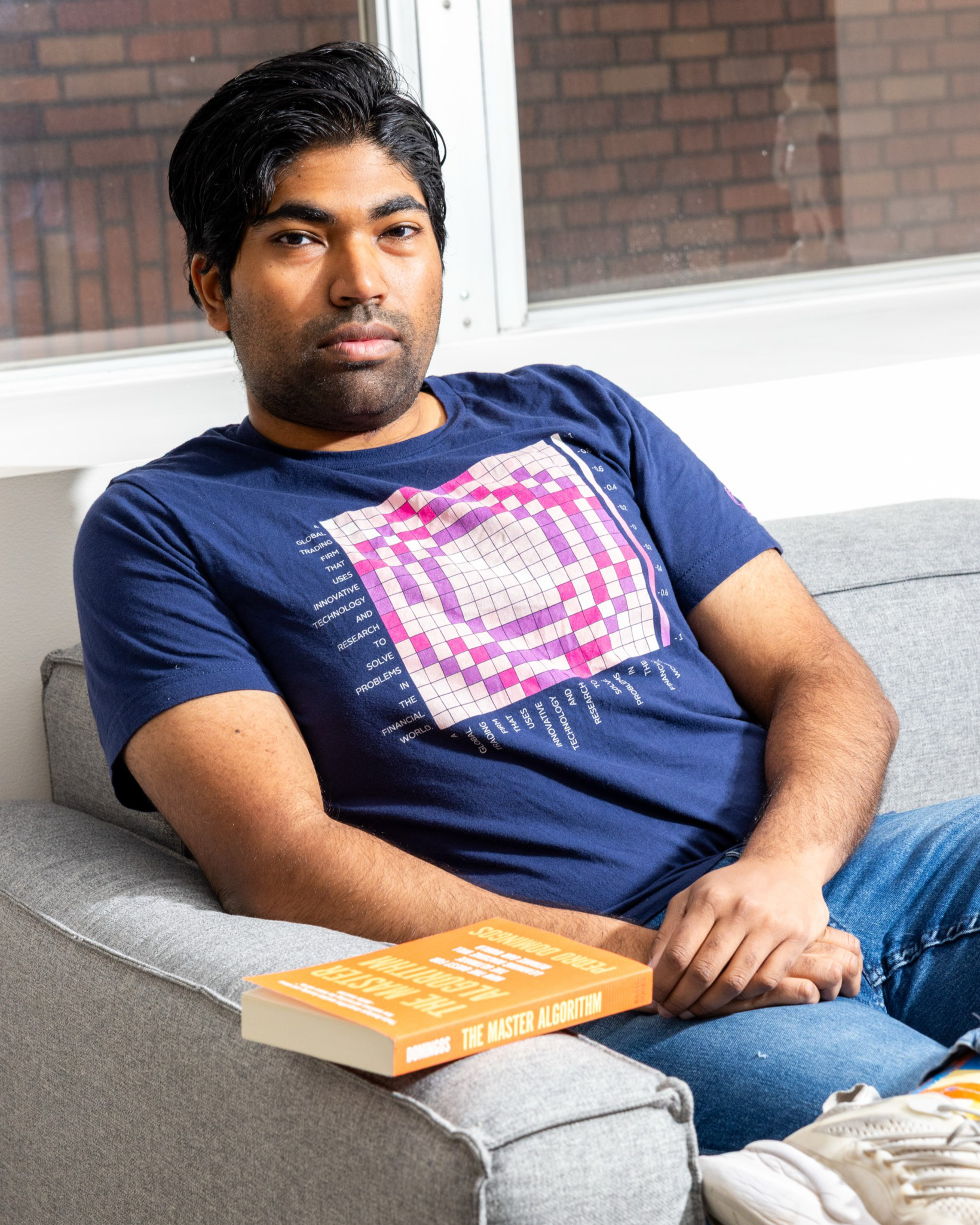 A man with dark hair sits on a gray couch wearing a blue graphic t-shirt and jeans, with a book titled “The Master Algorithm” resting on the armrest.