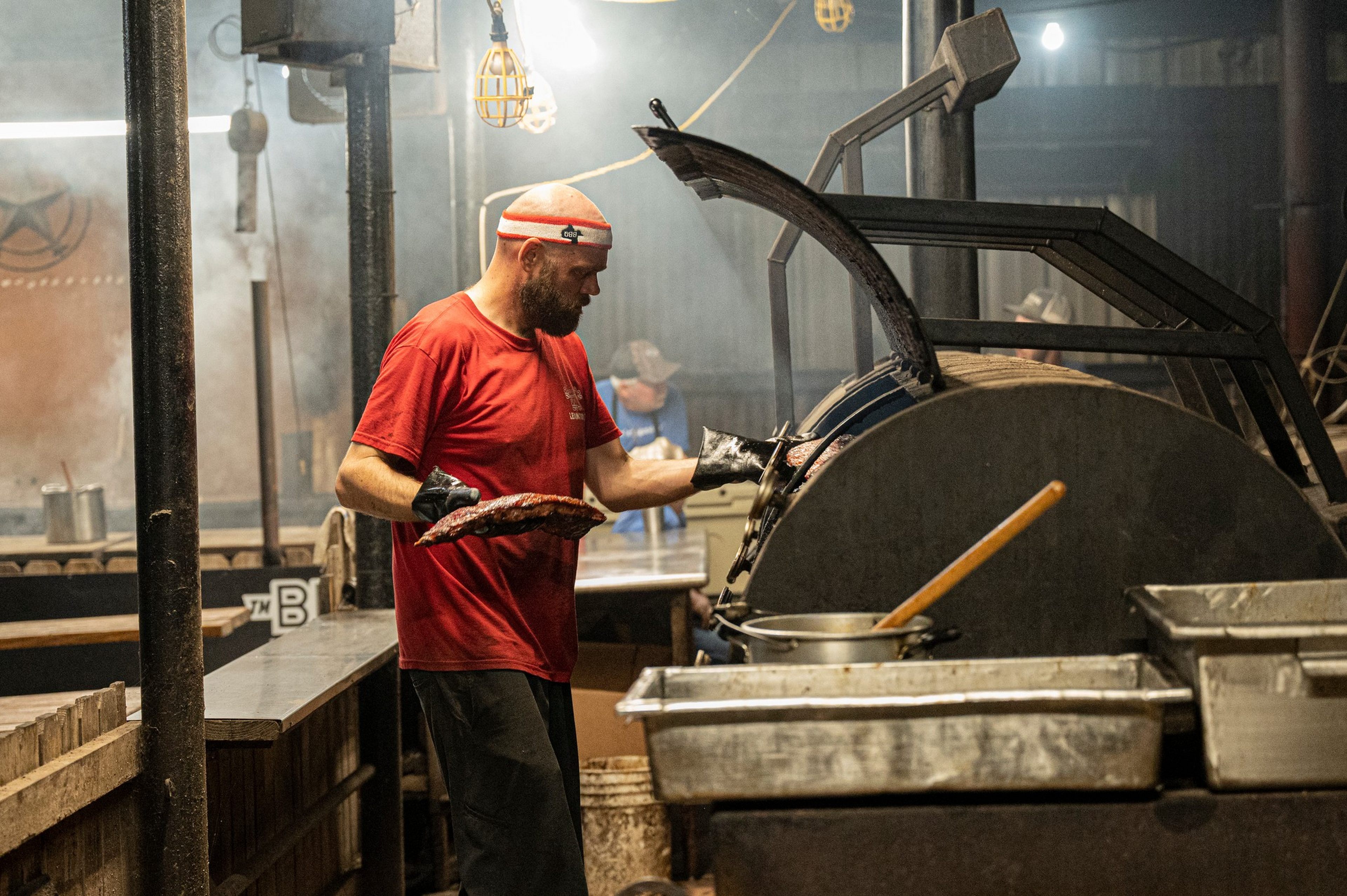 A man in a red shirt and headband uses black gloves to remove ribs from a large, open barbecue smoker in a rustic kitchen setting.