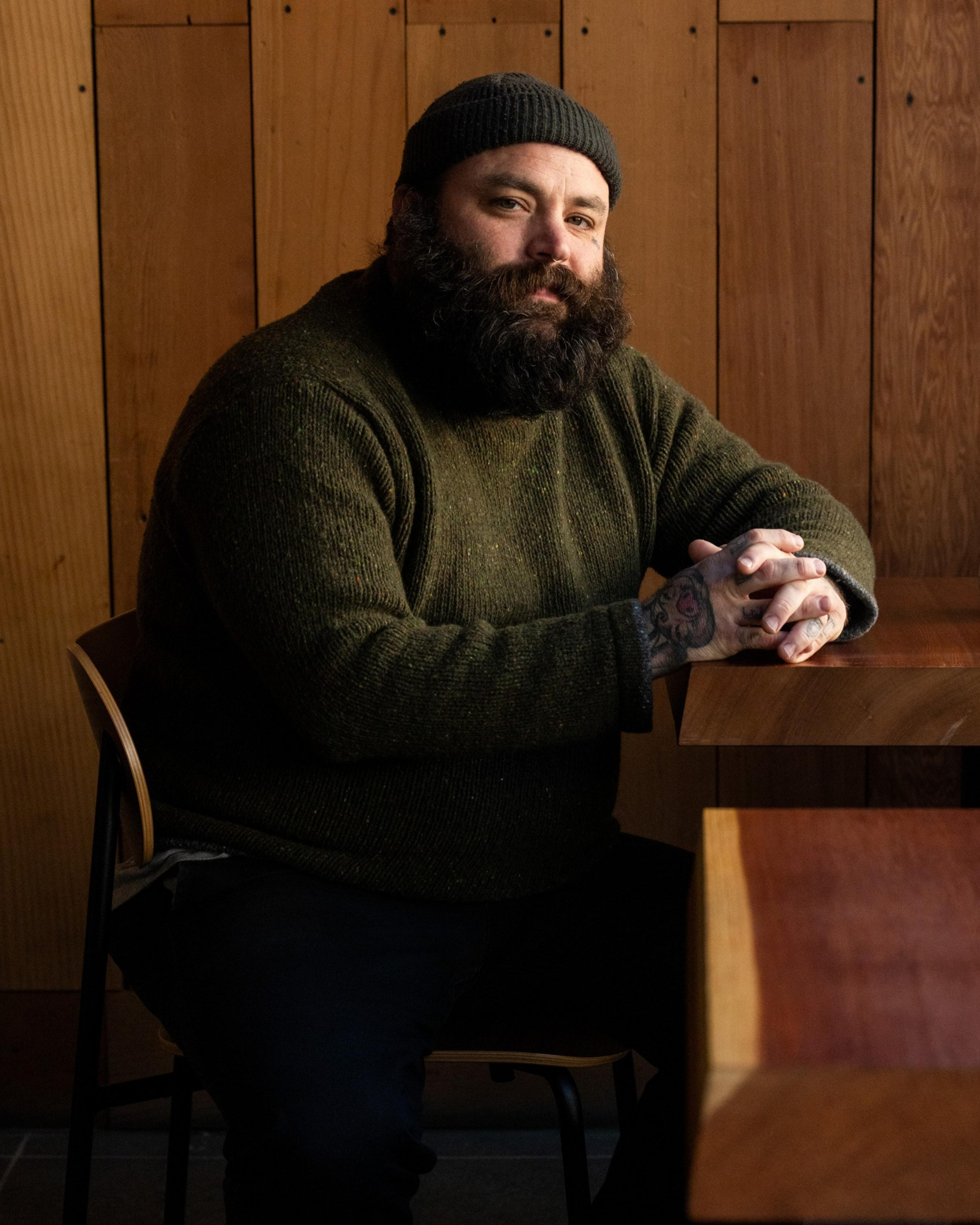 A bearded man in a green sweater and beanie sits at a wooden table, hands clasped. The background features wood paneling, creating a warm, rustic feel.