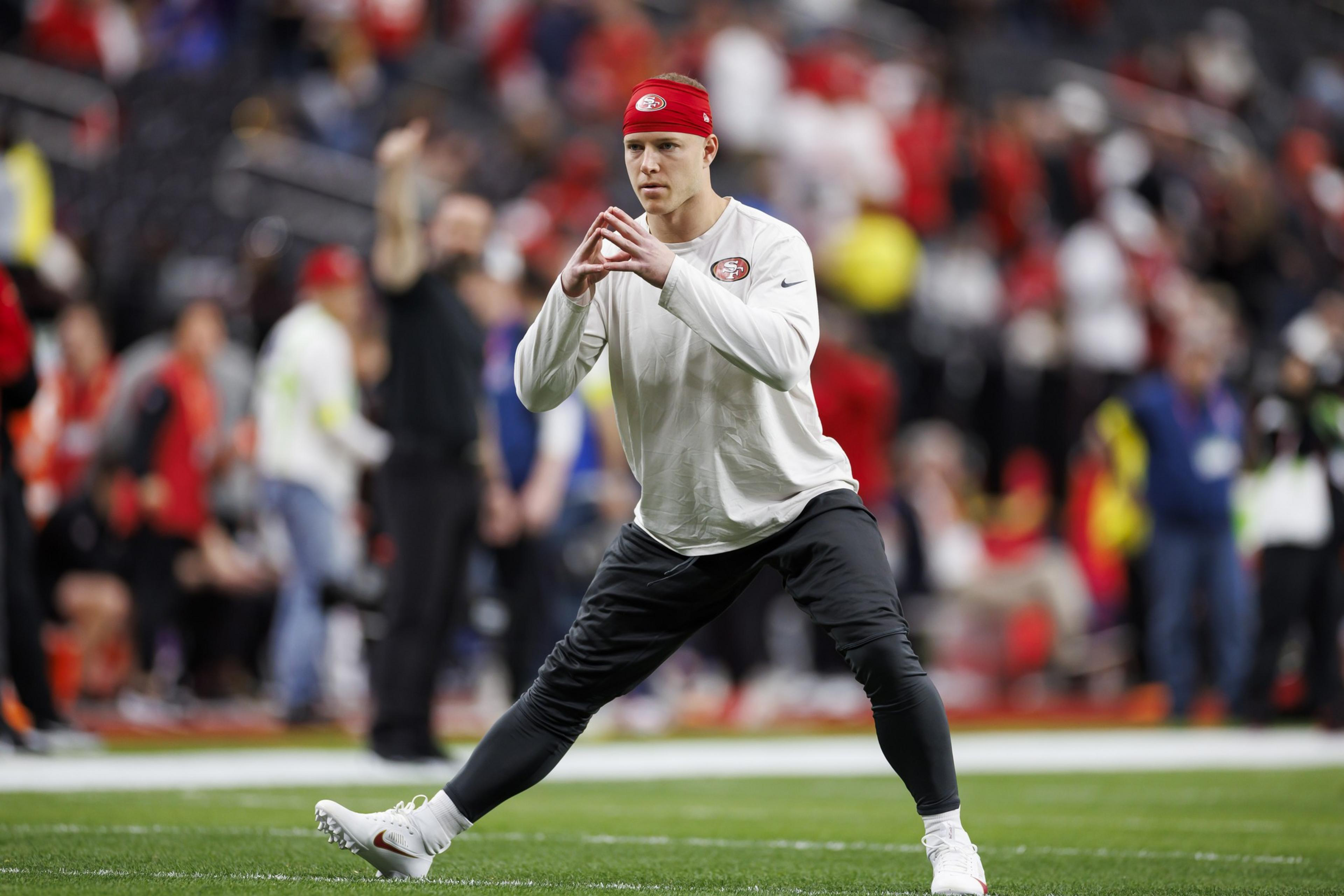 A man in a white long-sleeve shirt and red headband stretches on a football field. He's focusing, with hands clasped, while people in the background watch.