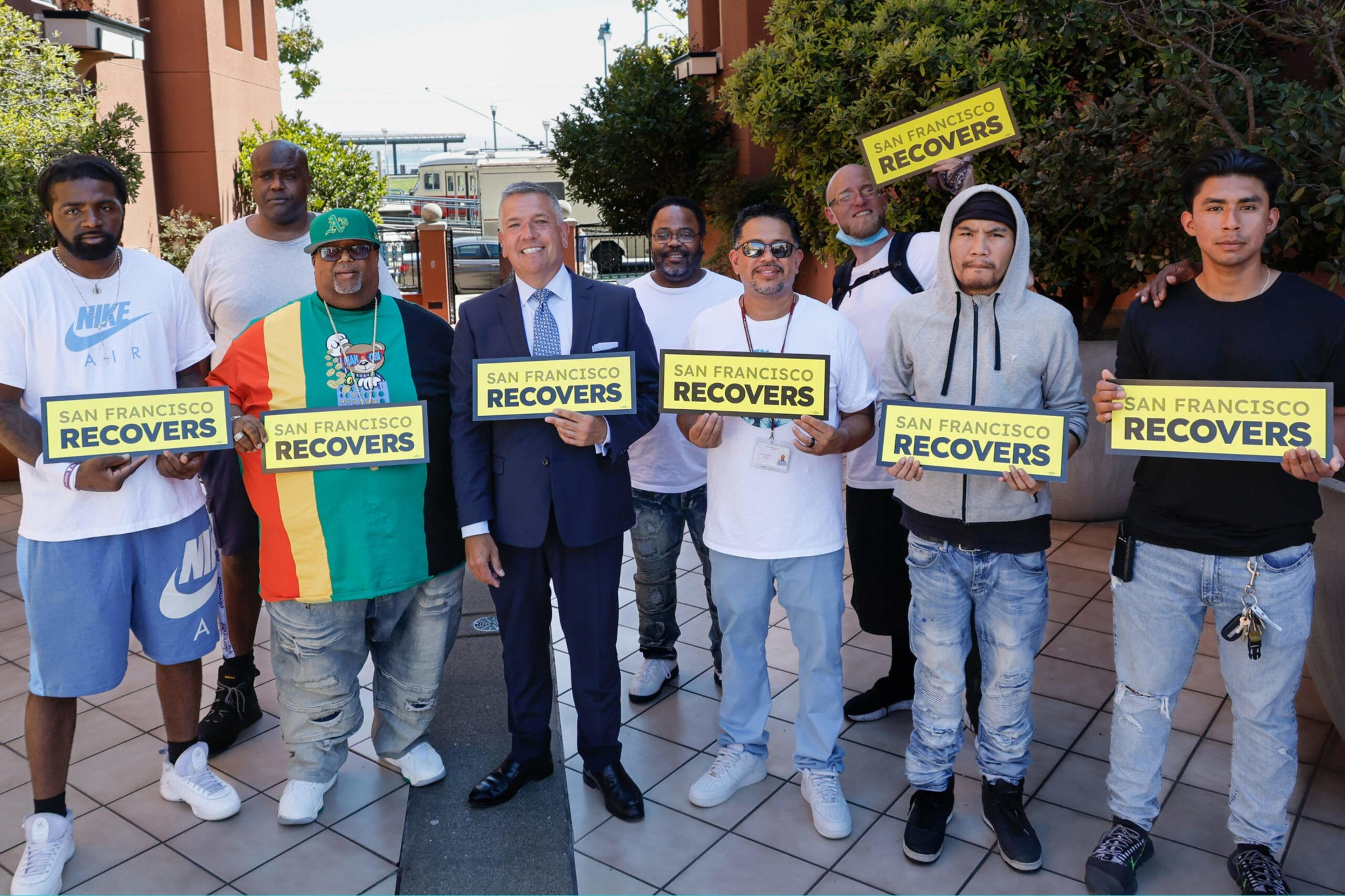 Nine men stand outdoors, smiling and holding yellow signs that say "San Francisco Recovers," with one man dressed in a suit and others in casual clothing.