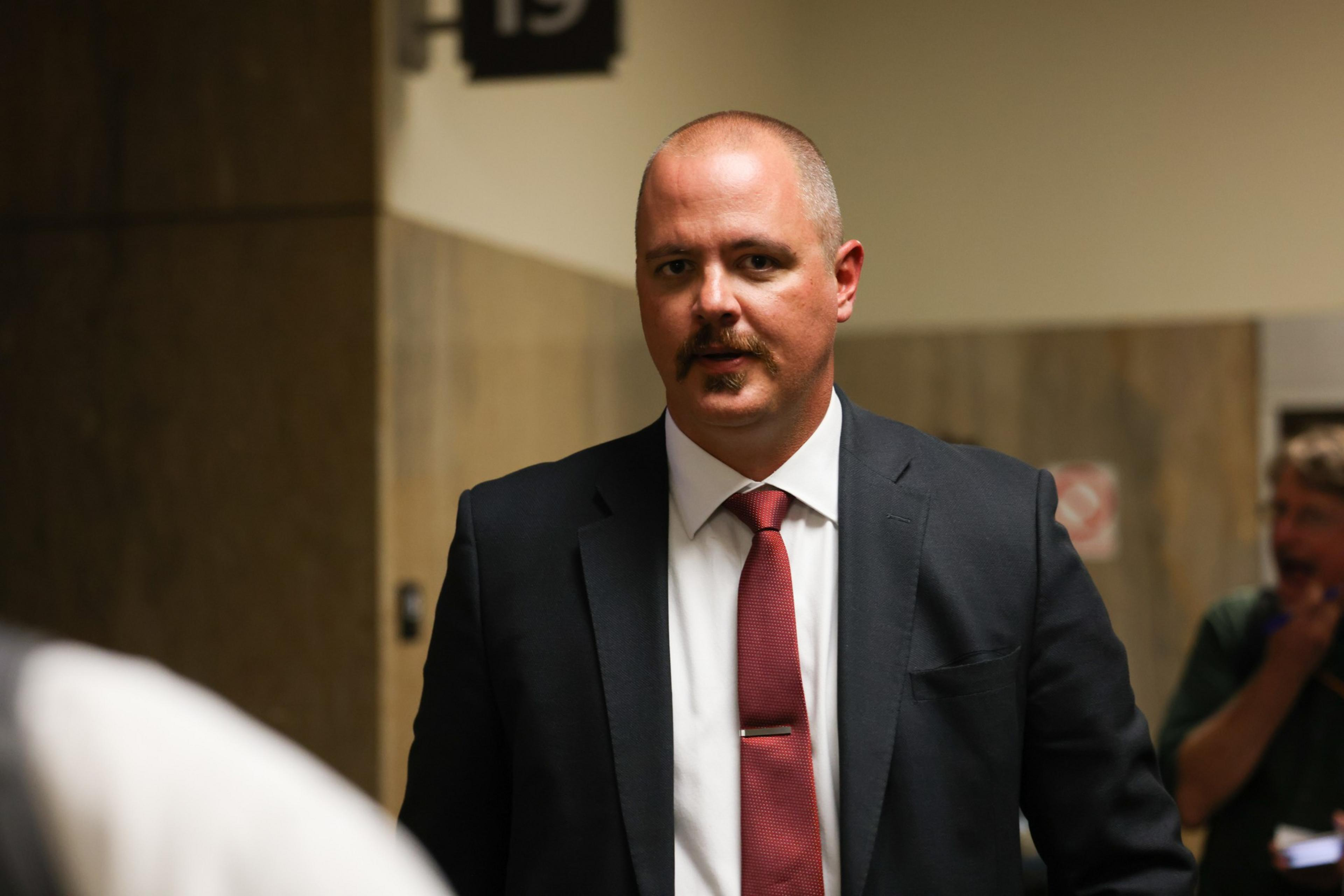 A man in a suit and red tie stands in a dimly lit hallway. He has a mustache and short hair. There's a blurred person in the background.