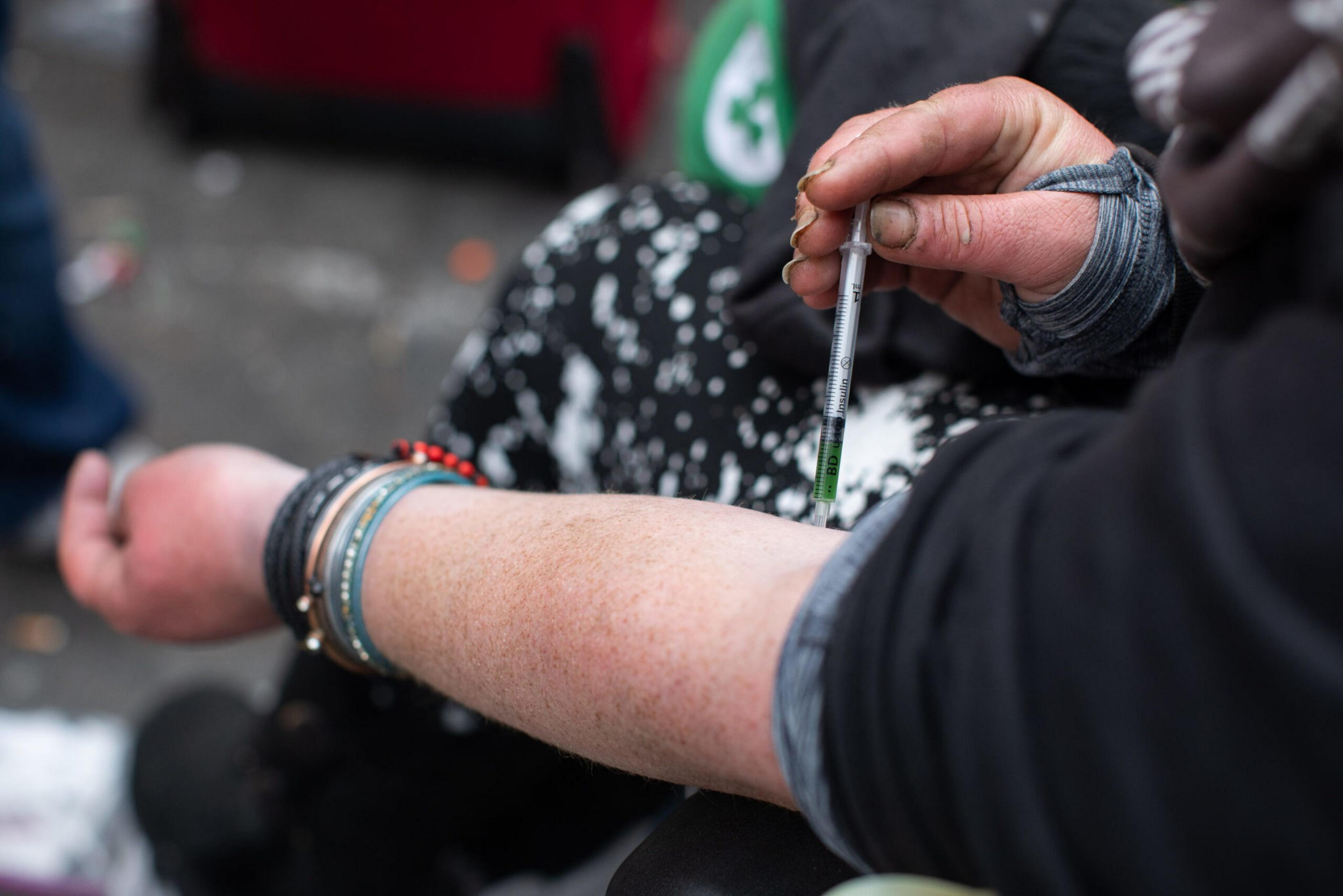 A person, wearing multiple bracelets and a black outfit, injects a substance into their forearm using a syringe. The background is slightly blurred.