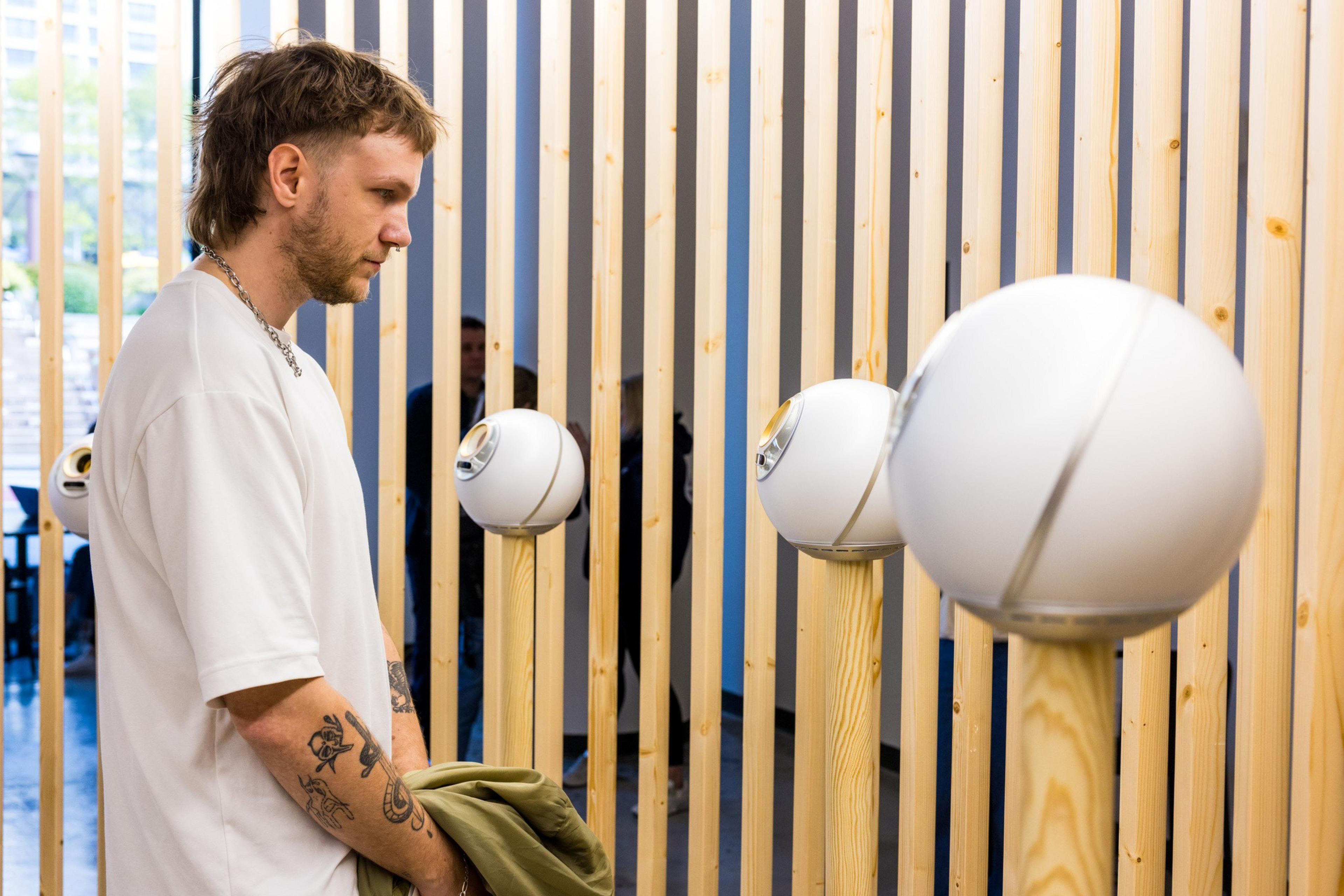 A person with tattoos and a mullet hairstyle stands near a row of modern speakers mounted on wooden posts, with vertical slats in the background.