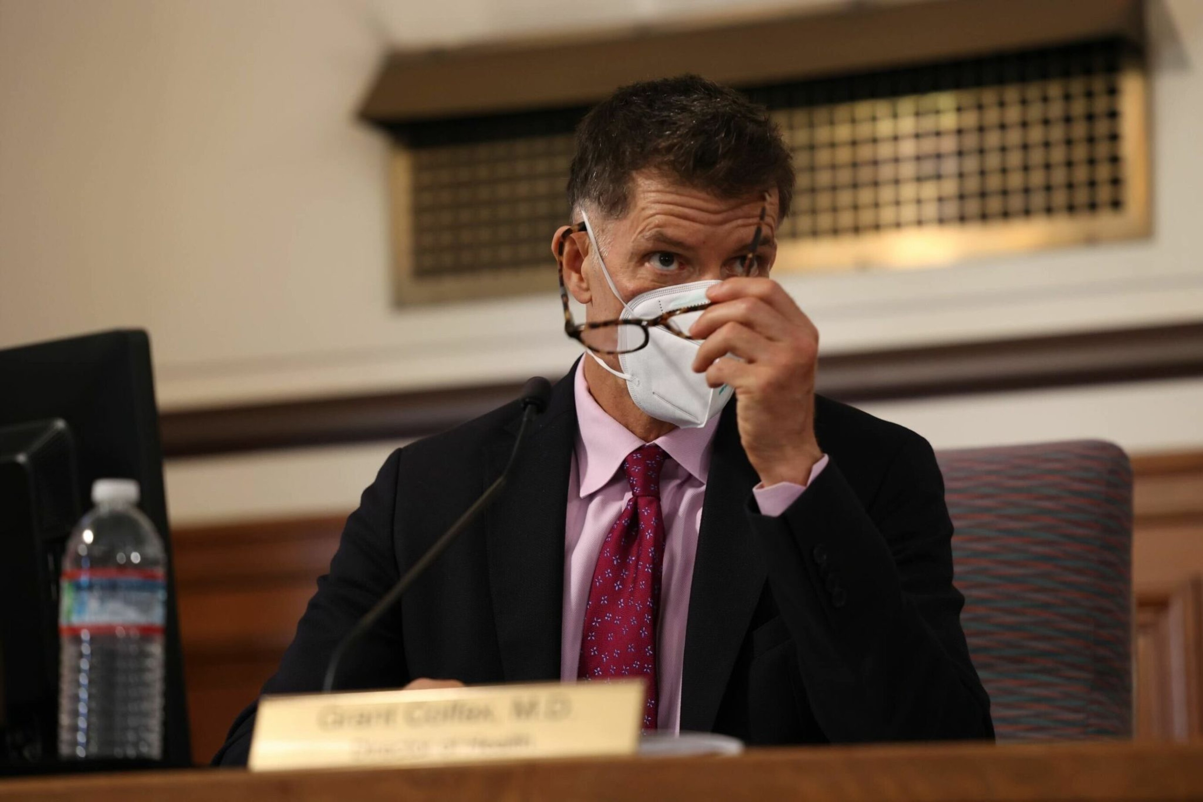 A man in a suit and red tie wears a white mask, holding eyeglasses near his face while sitting behind a nameplate and microphone.