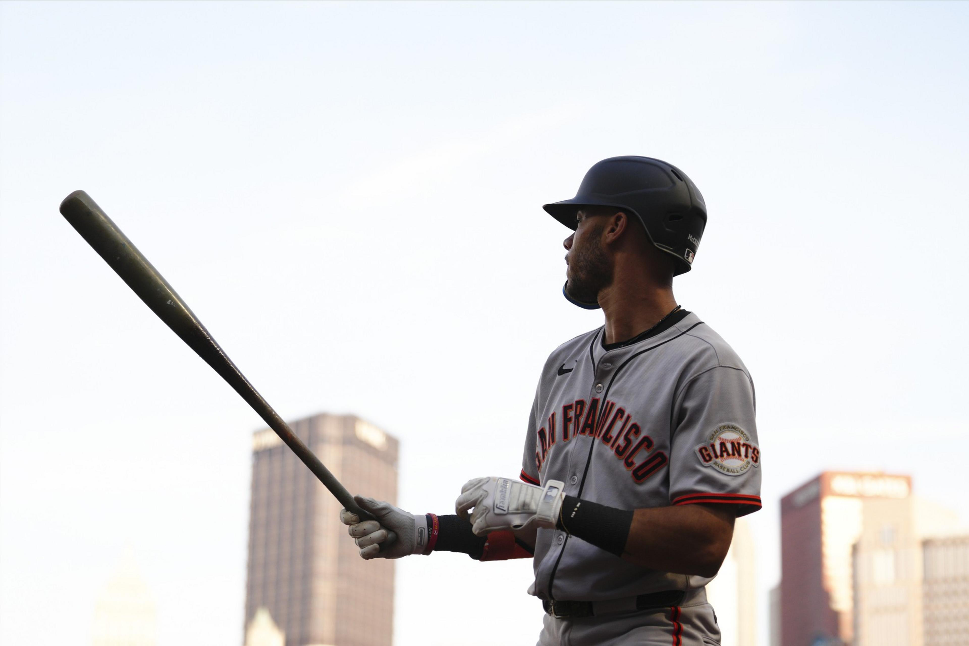 A baseball player in a San Francisco Giants uniform holds a bat, looking off to the side with city buildings blurred in the background.