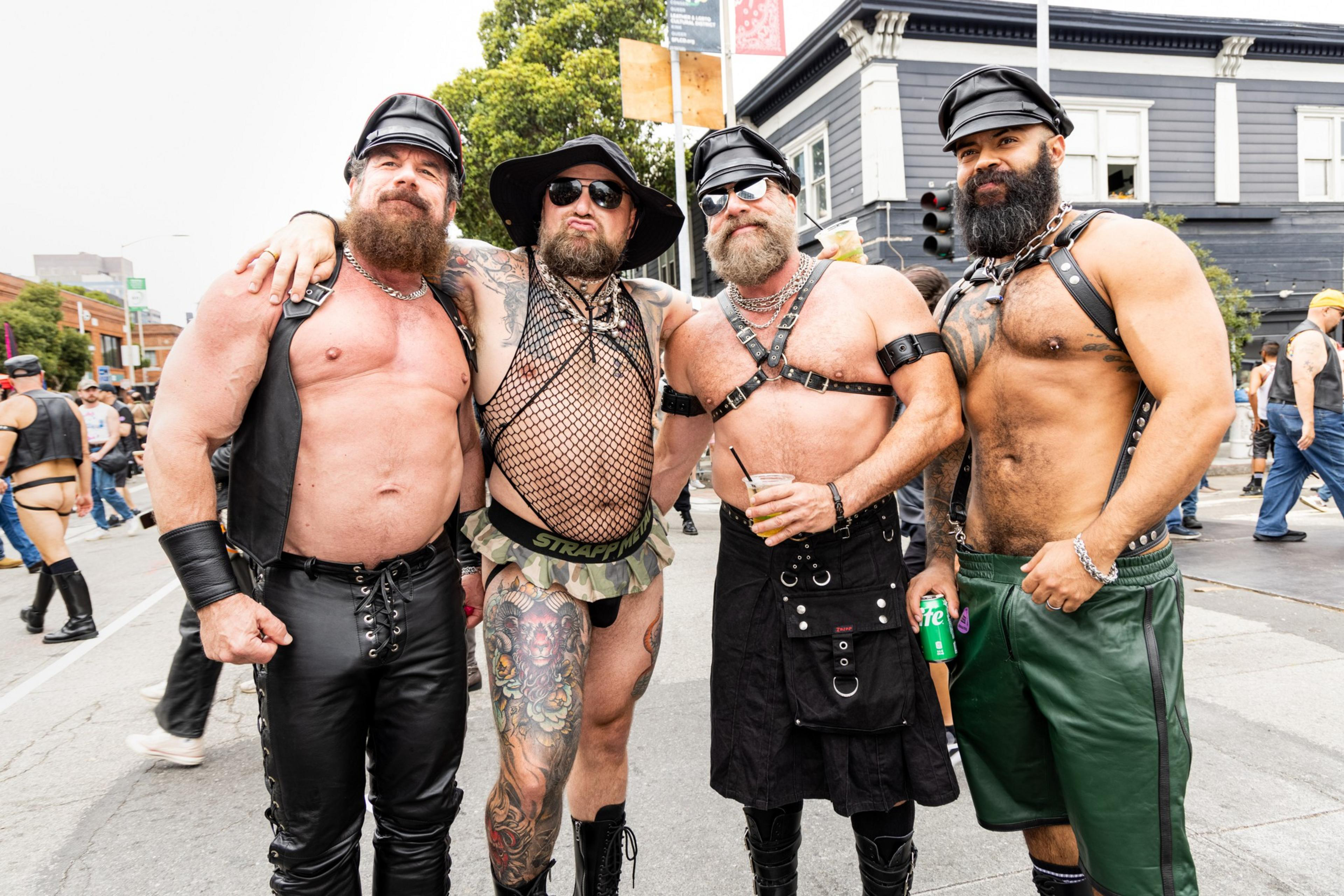 Four muscular men with beards wear leather and harness outfits, standing close together on a street during an outdoor event.