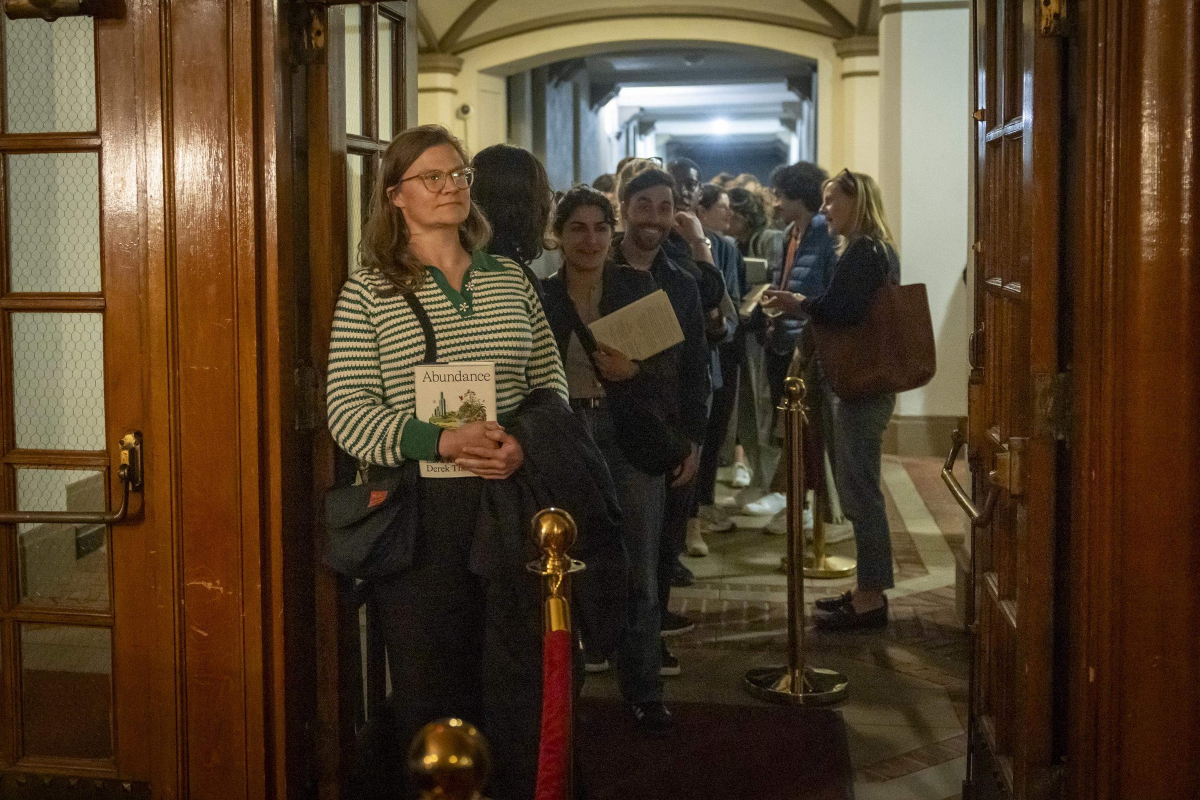 A woman in a striped sweater holds a book titled &quot;Abundance&quot; in a line of people waiting inside a building, with a red rope barrier beside them.