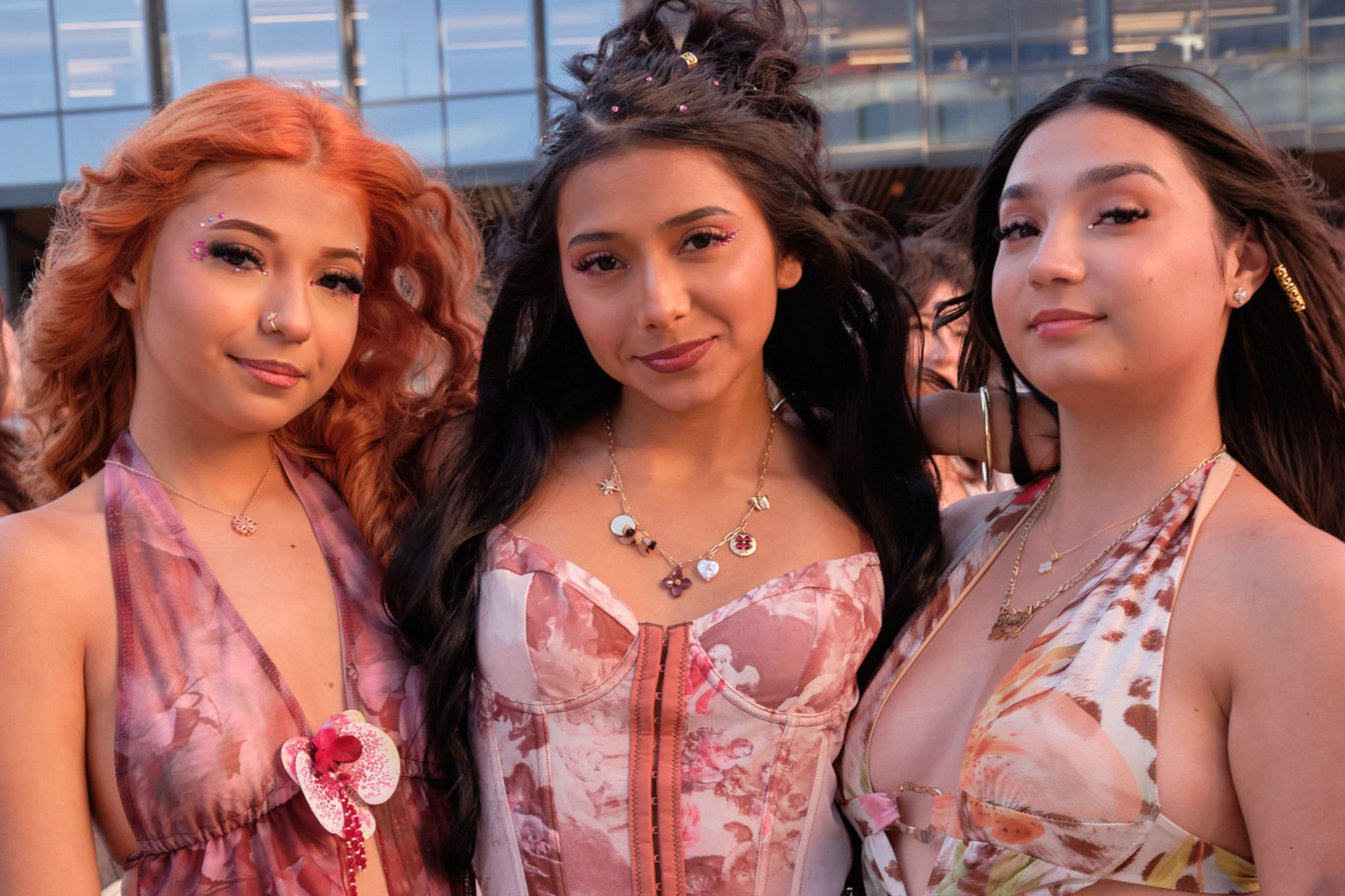 Three young women stand close together outdoors, wearing floral patterned dresses and skirts in shades of pink and peach, accessorized with jewelry and light makeup.