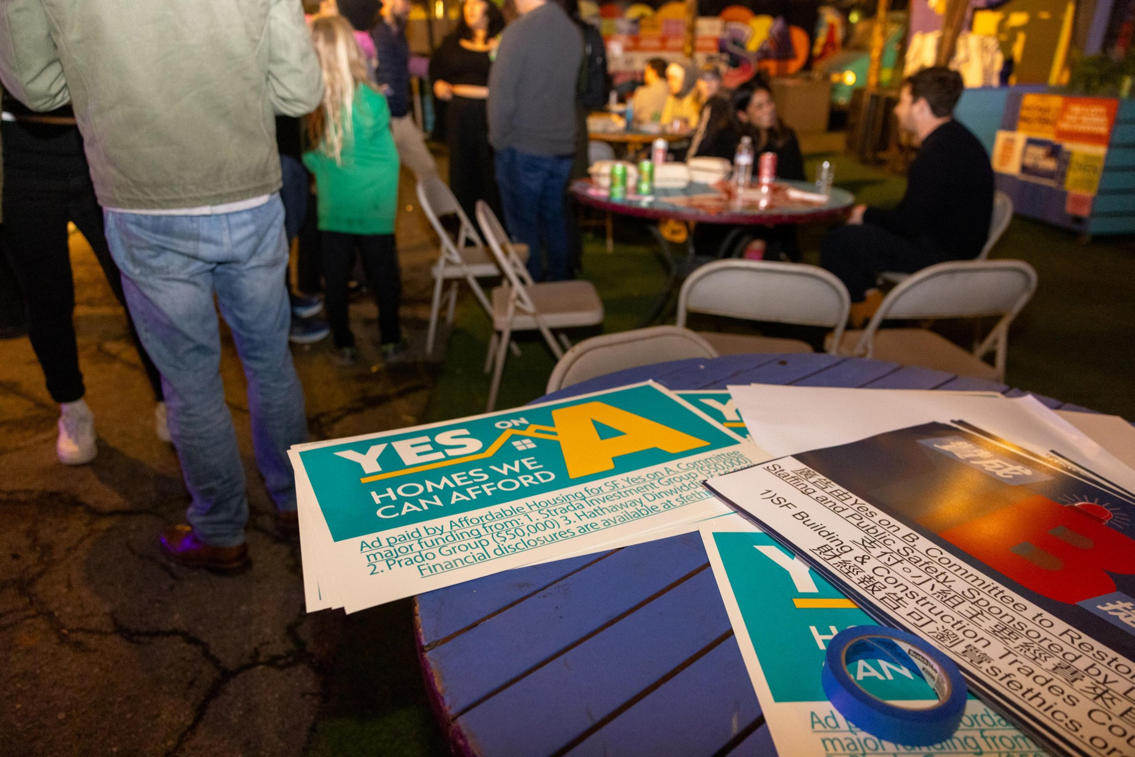 The image shows a social gathering with people standing and sitting around tables. On a table in the foreground, there are campaign signs reading &quot;Yes on A&quot; and &quot;Yes on B.&quot;