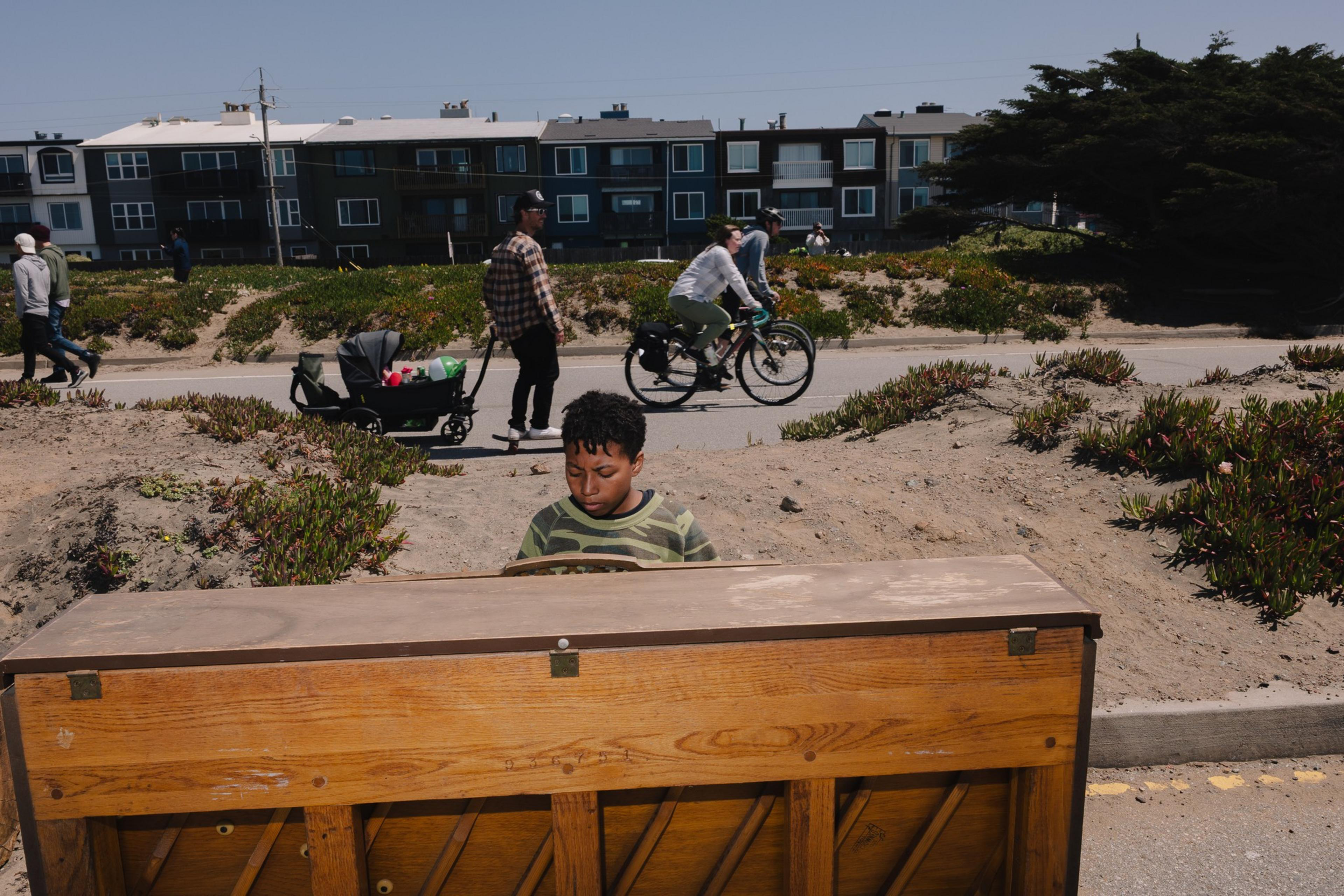 A young boy plays an upright piano outdoors near a path, with people walking and cycling nearby. Behind them are low-rise buildings and trees under a clear sky.