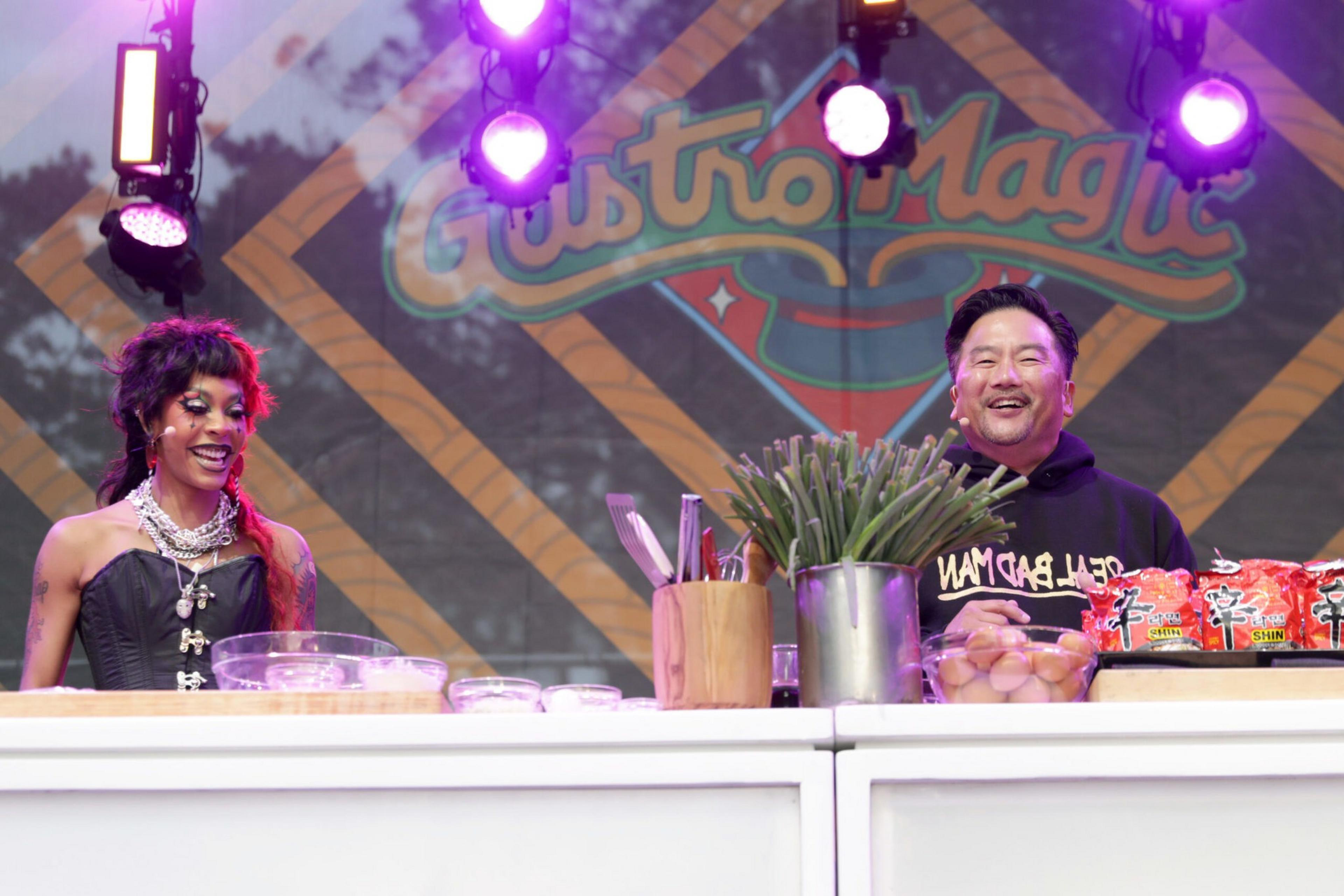 Two people stand behind a cooking counter with ingredients like eggs, green vegetables, and ramen noodles, under purple stage lights with a "Gusto Magic" sign.
