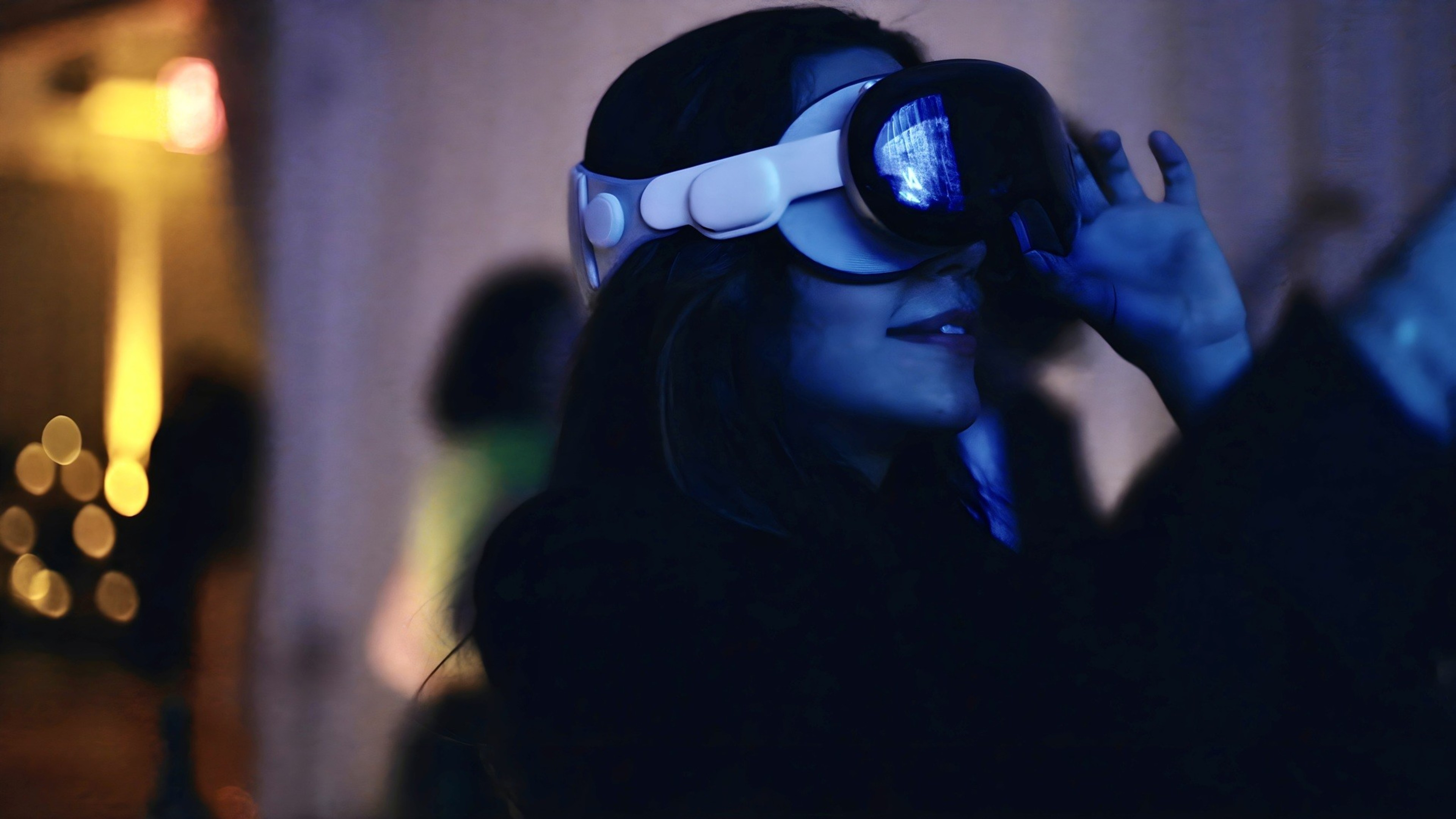 A woman wears an Apple Vision Pro headset, illuminated by glowing blue light, at a party.