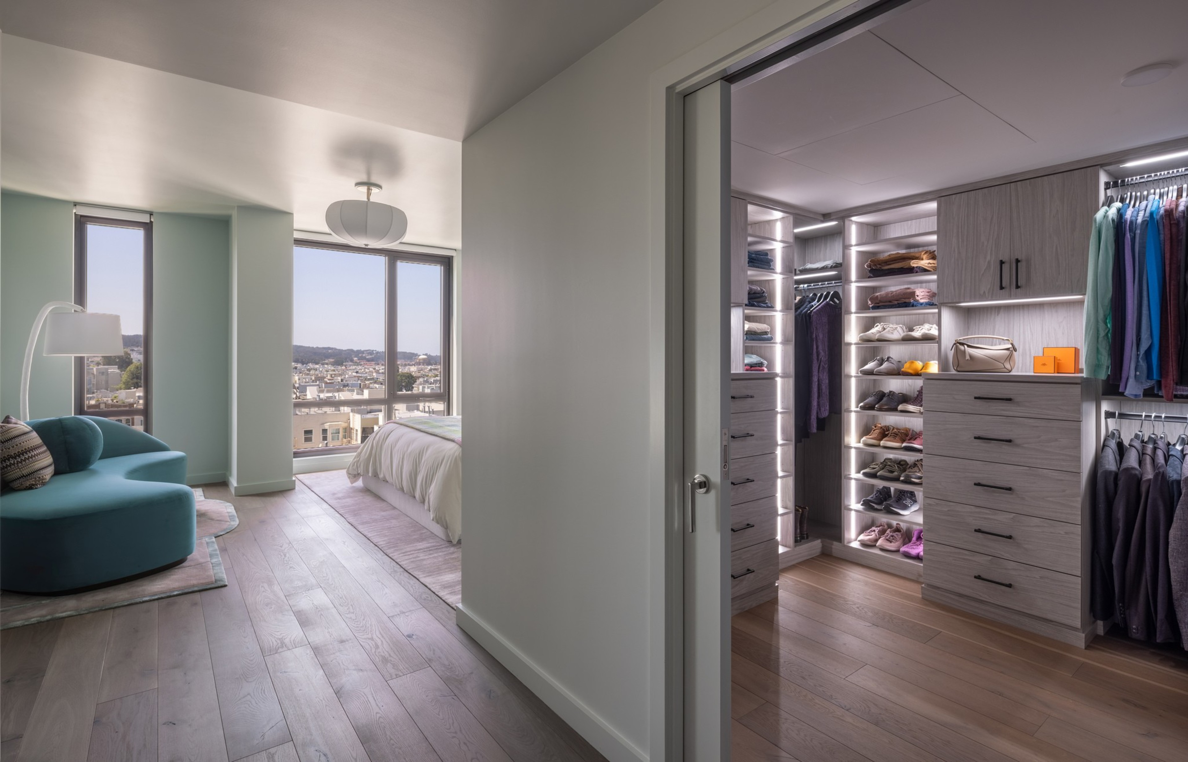 A bedroom with a blue armchair and floor lamp leads to a large walk-in closet with shelves, drawers, and hanging clothes illuminated by soft lighting.
