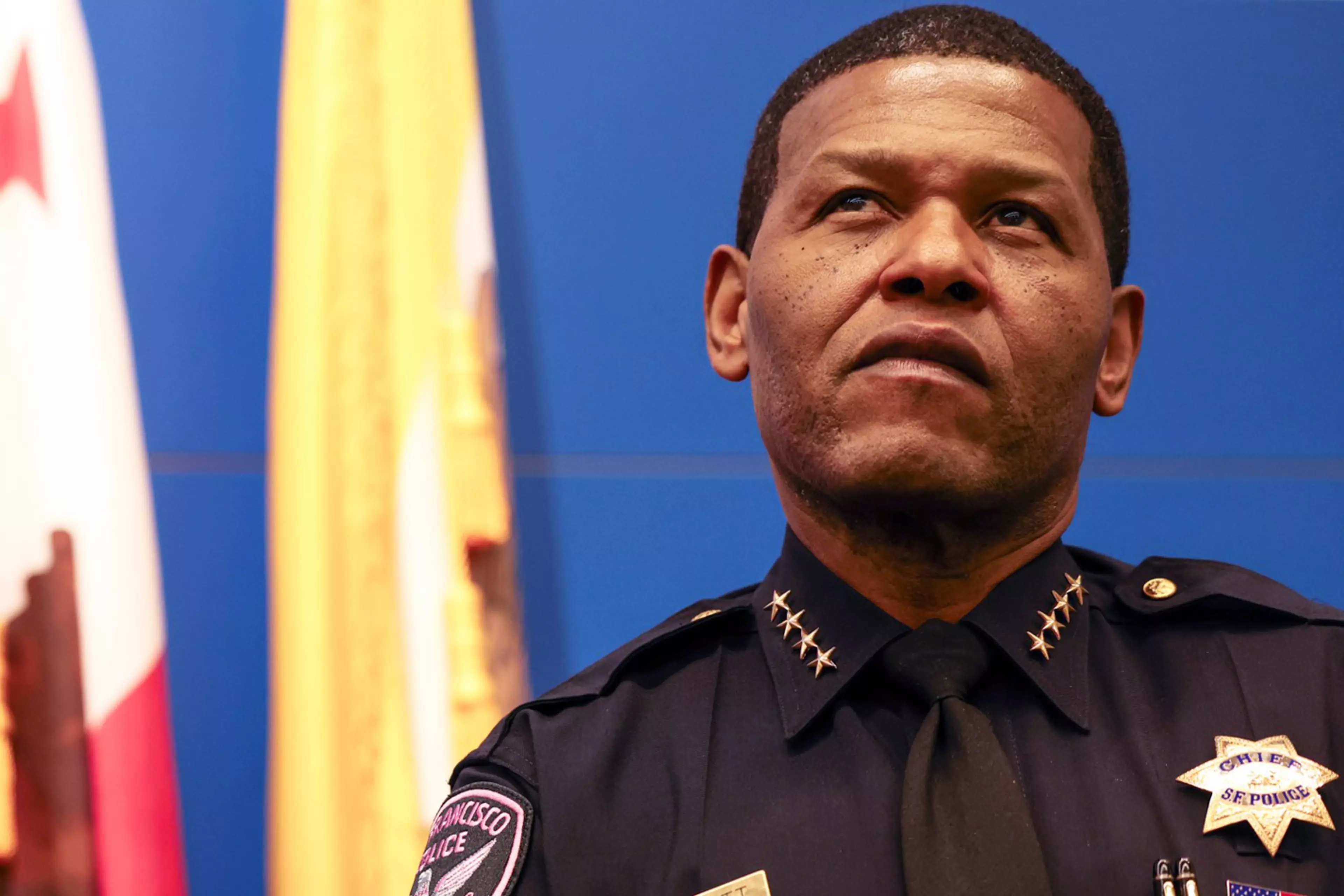A serious police chief in uniform with gold stars on his collar and badges on his chest stands before blurred flags.