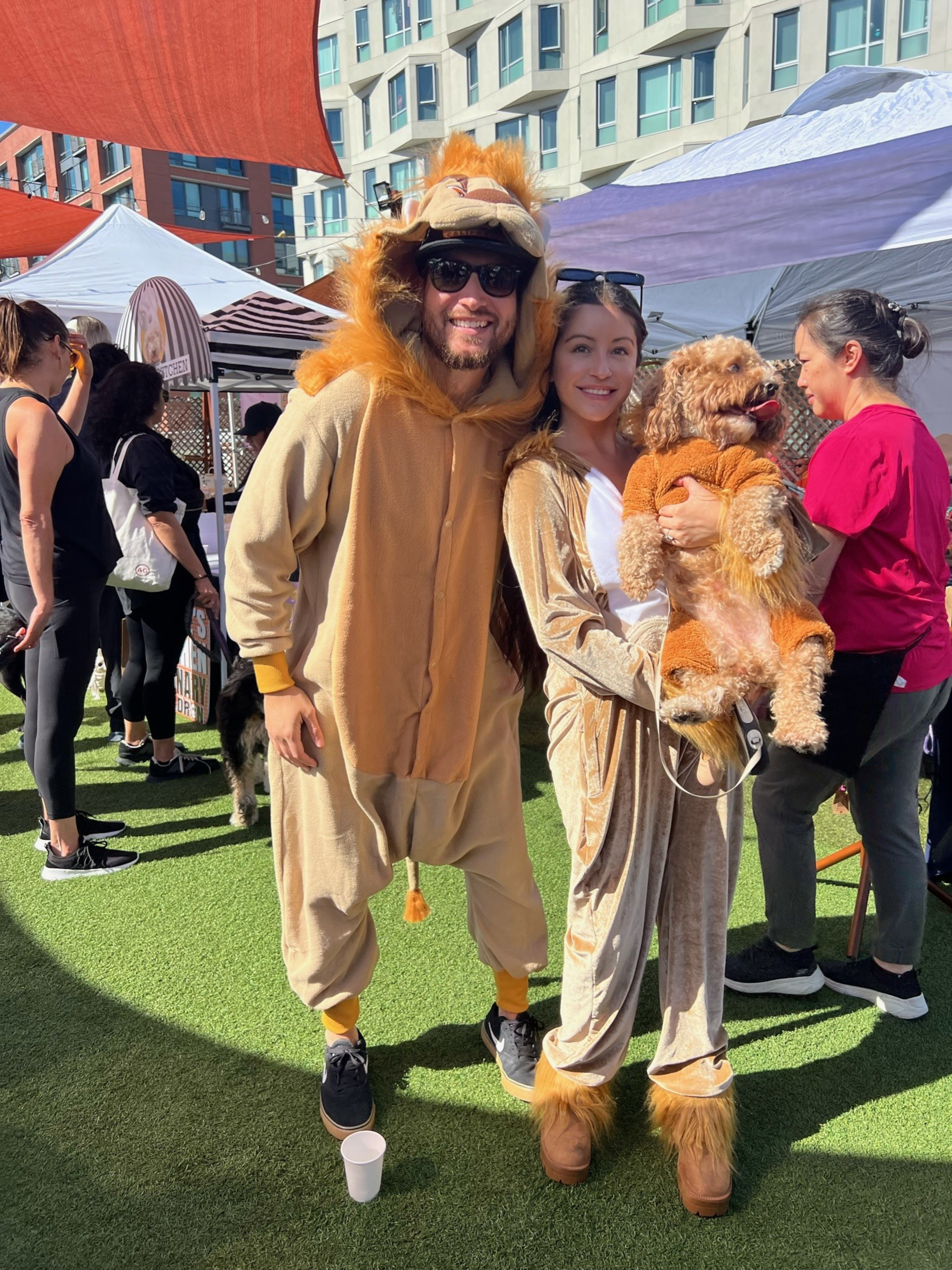 A man and woman wear matching lion costumes, the woman holds a small dog also dressed in a lion outfit, all smiling outdoors at a sunny event.