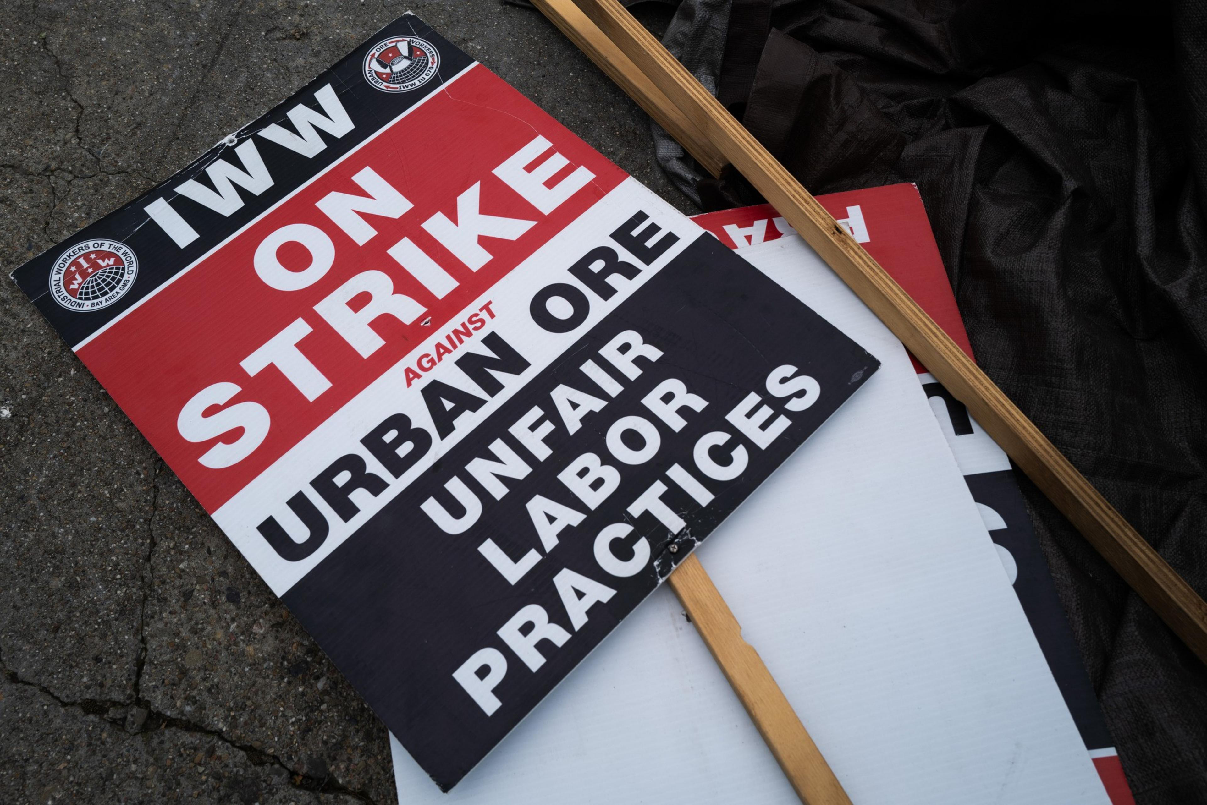 A protest sign on the ground reads &quot;IWW ON STRIKE against Urban Ore UNFAIR LABOR PRACTICES&quot; with bold red, black, and white colors.