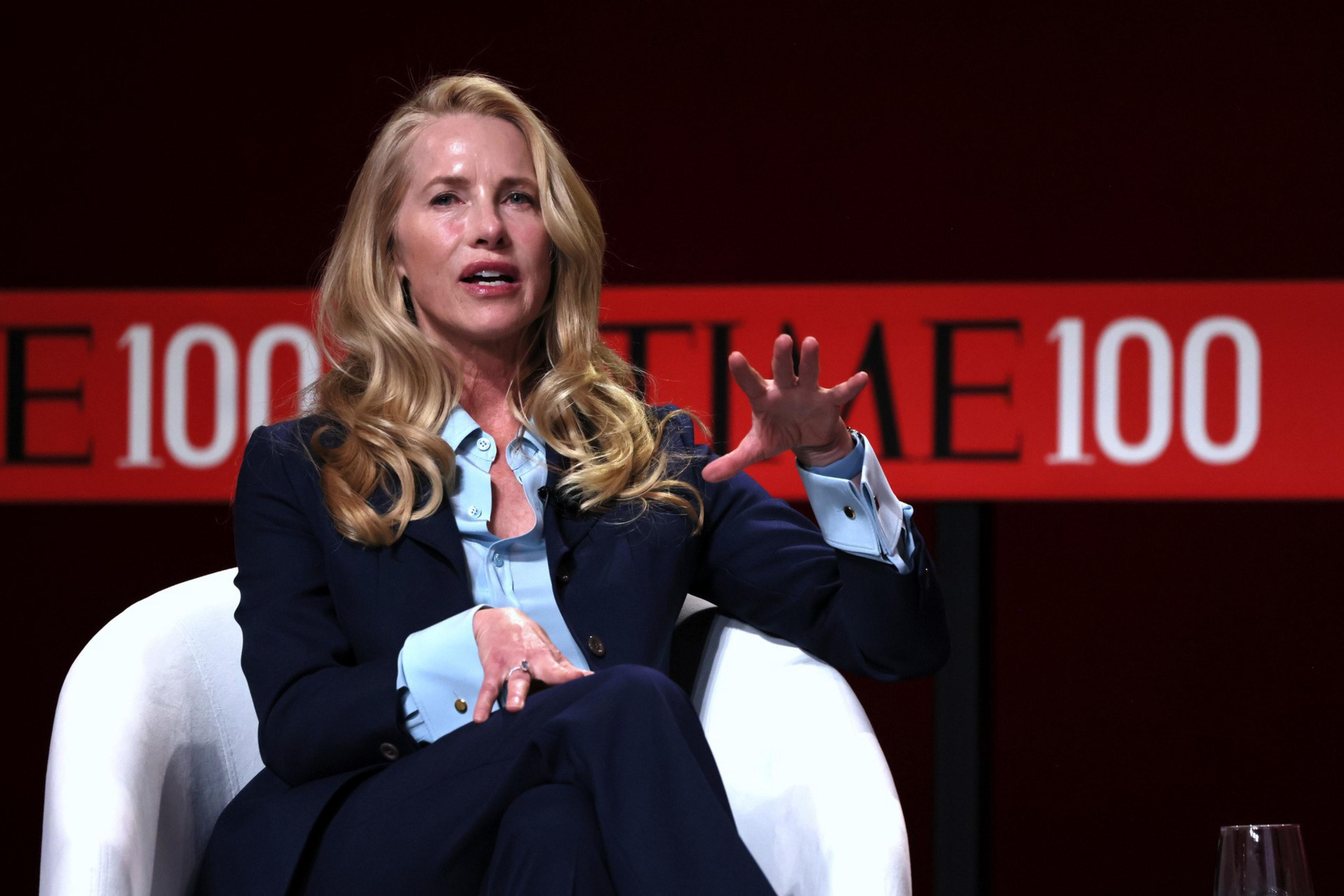 A woman with long blonde hair wearing a navy suit and blue shirt speaks while seated in a white chair, gesturing with her hand. A "TIME 100" banner is in the background.
